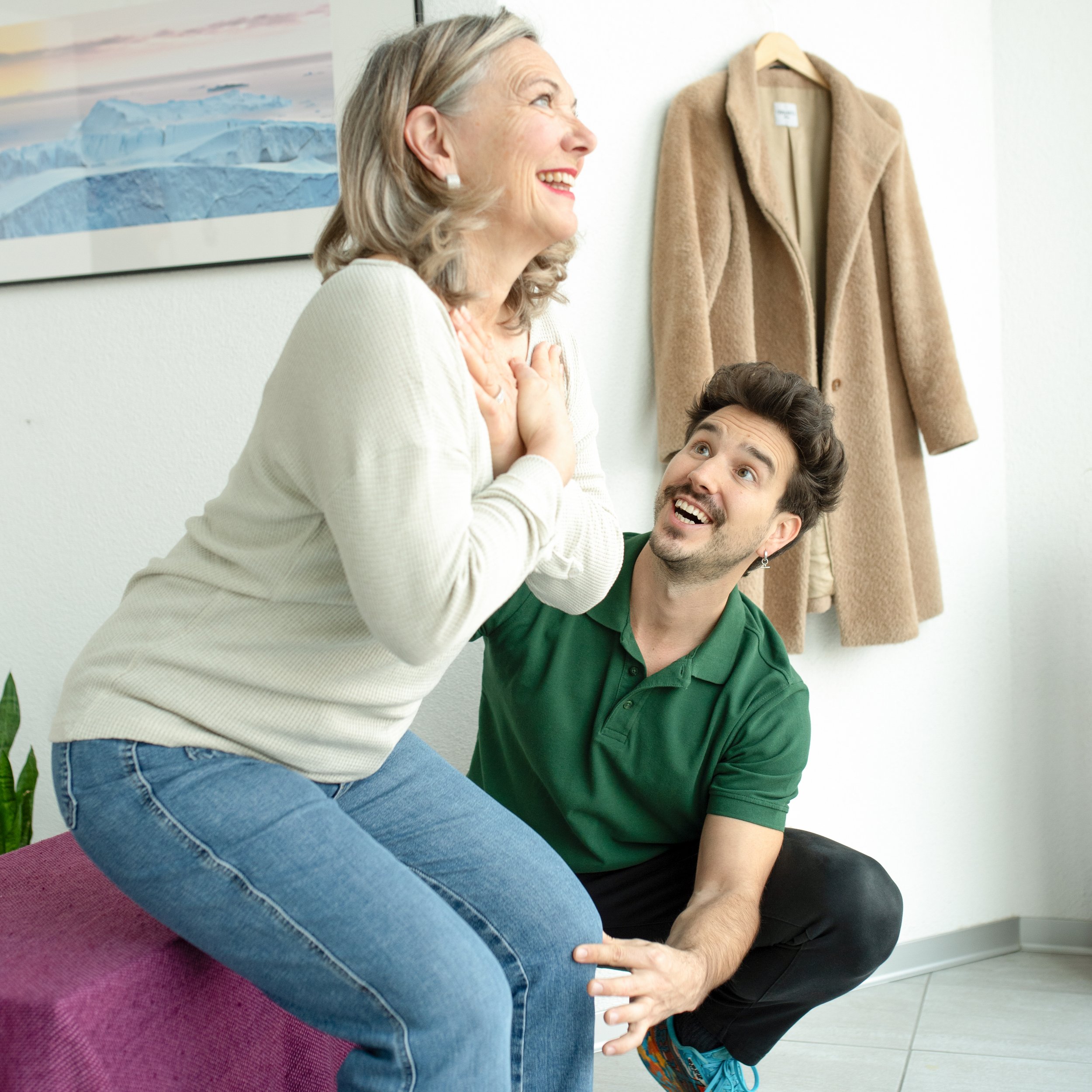 Ein älterer Frau sitzt auf einem pinkfarbenen Hocker, während ein junger Mann sie unterstützend hält und mit ihr lacht. Im Hintergrund hängt ein beiger Mantel an der Wand, und auf einem Bild ist eine Gletscherlandschaft zu sehen.