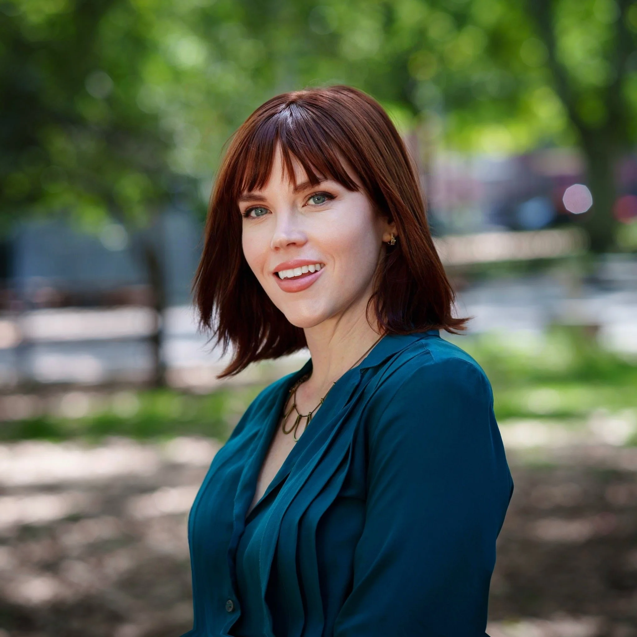 Portrait of a woman with shoulder-length red hair, smiling, wearing a teal blouse and gold jewelry, outdoors with green trees in the background.