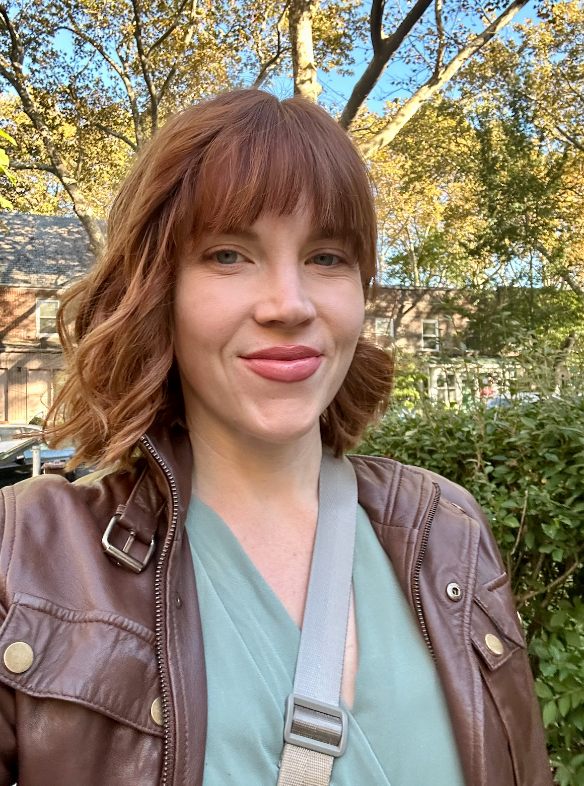 A woman with red hair and light skin smiling outdoors, wearing a brown leather jacket and a light green top, with trees and buildings in the background on a sunny day.