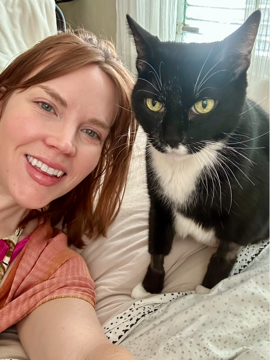 A woman with red hair and a big smile taking a selfie with a black and white tuxedo cat on a bed or couch.