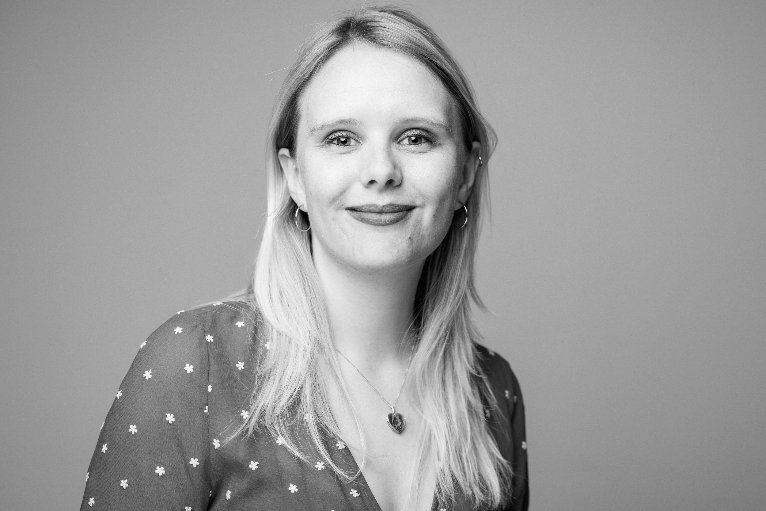 Black-and-white portrait of a smiling woman with light-colored hair, wearing hoop earrings, a necklace with a heart-shaped pendant, and a dark top with small floral patterns, against a plain background.