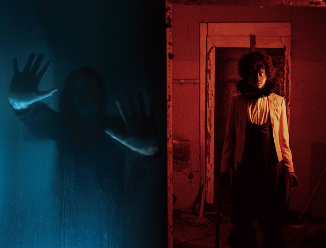 A person with curly hair standing in a dimly lit room, wearing a beige blazer and black clothing, illuminated by red light.