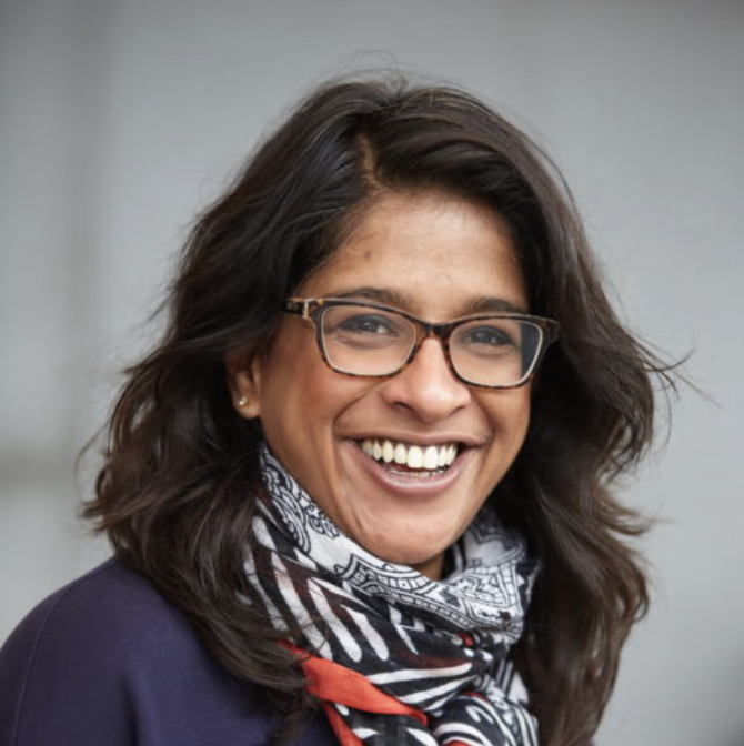 A woman with medium skin tone, glasses, and dark wavy hair smiling at the camera, wearing a patterned scarf and a dark top.