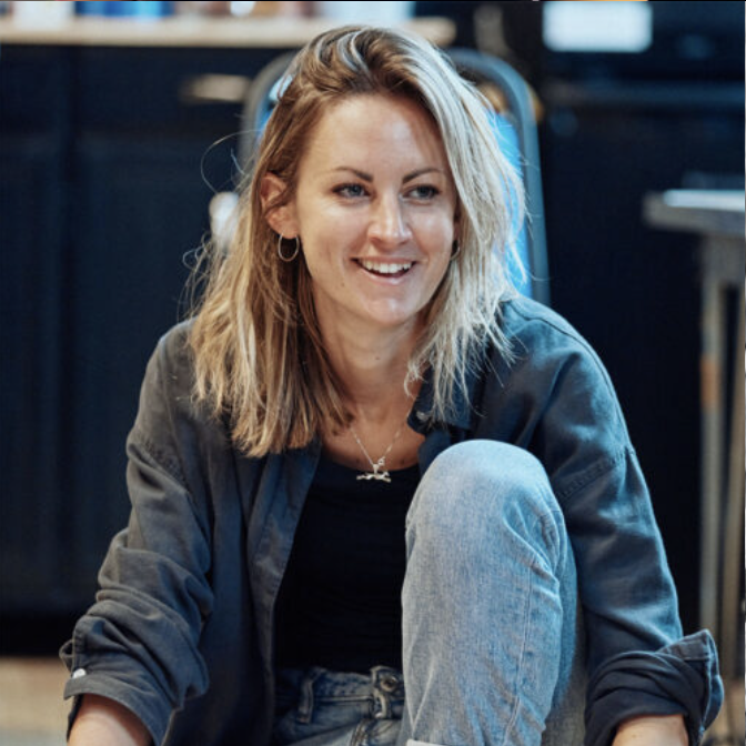 Smiling woman with blonde hair sitting on the floor in an indoor setting, wearing a dark jacket and jeans.
