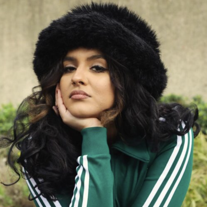 Woman with dark curly hair wearing a black fur hat, green track jacket with white stripes, sitting outdoors with a neutral expression.