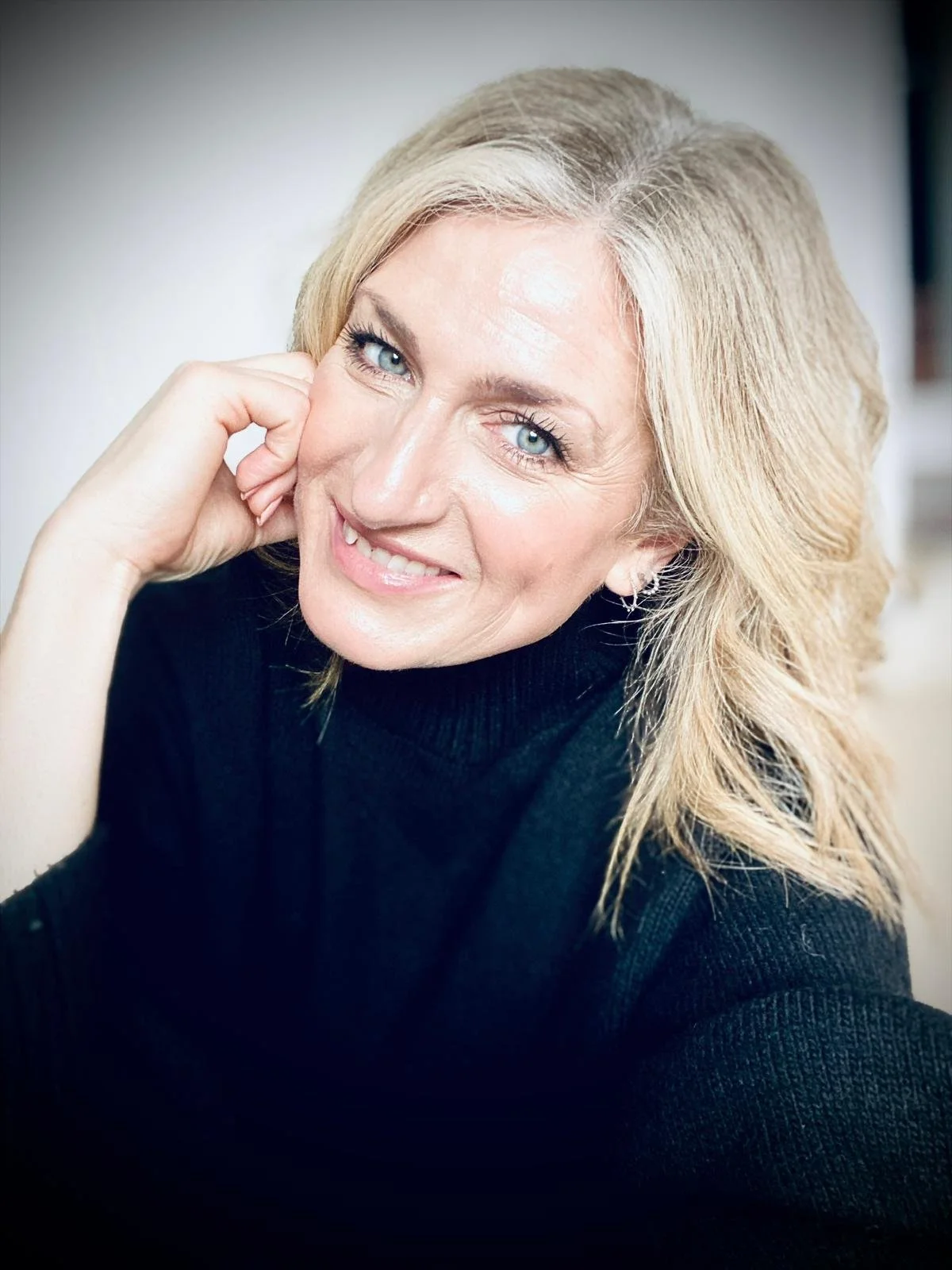 A smiling woman with blonde hair and blue eyes, wearing a black turtleneck, resting her face on her hand.