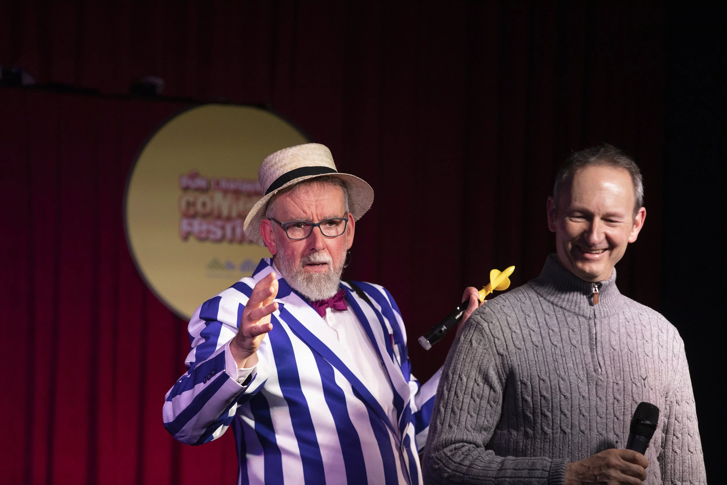 Two men on stage, one dressed in a striped suit with a hat and glasses, the other in a gray sweater holding a microphone, with a red curtain and a circular sign in the background.