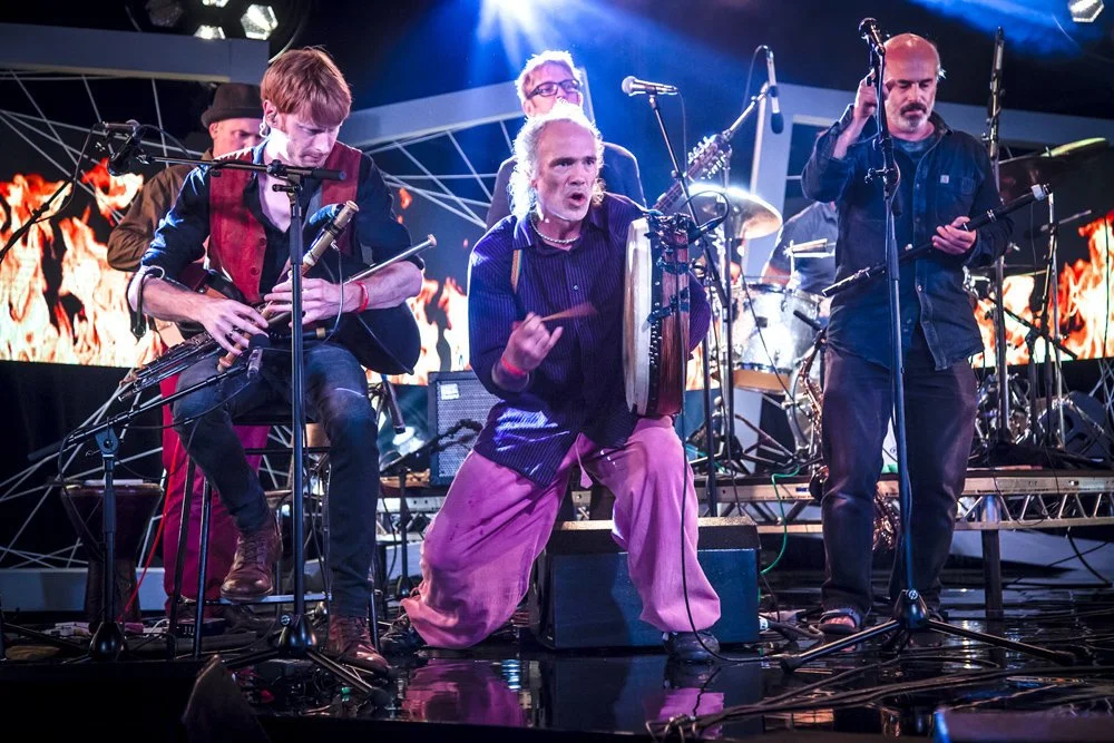 A band performing live on stage with multiple musicians playing traditional instruments, including drums and a tambourine, with a vibrant digital backdrop.