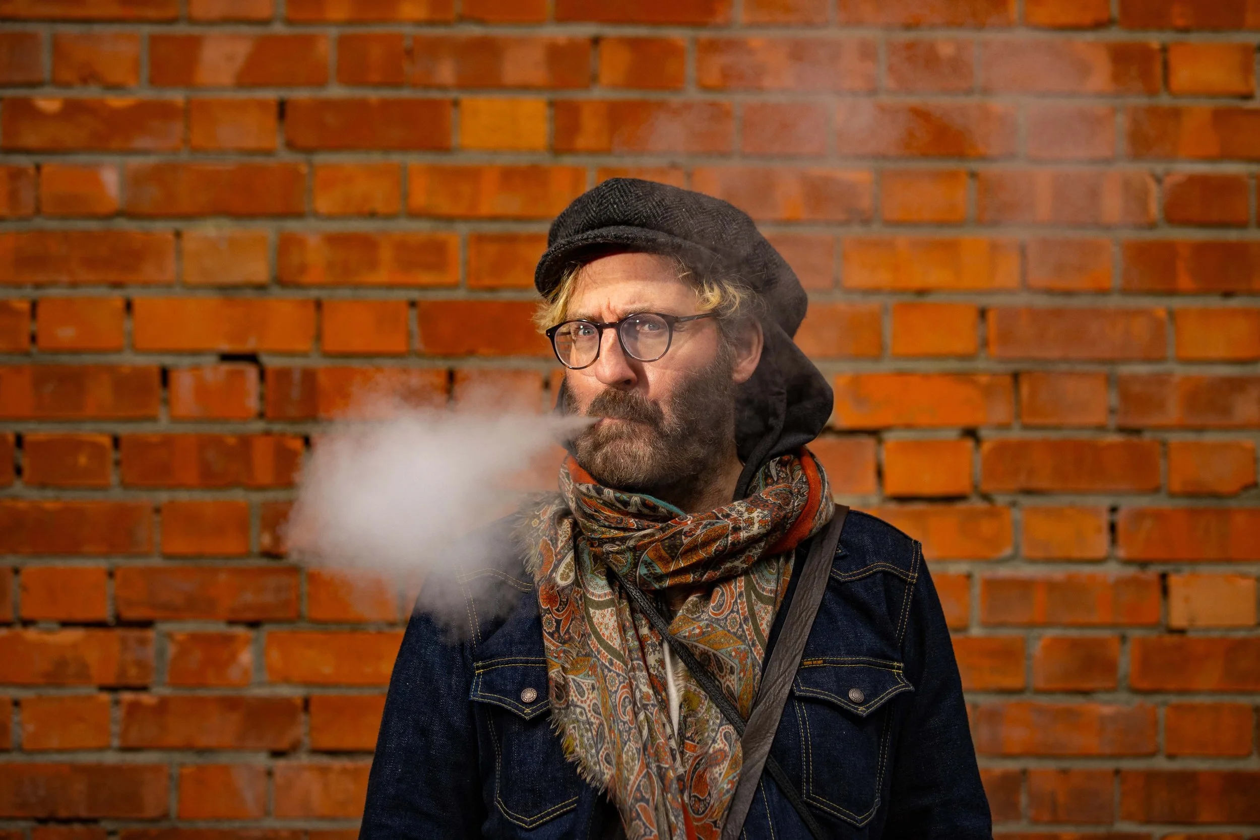 A man with glasses and a beard, wearing a hat and scarf, exhales vapor while standing against a brick wall.