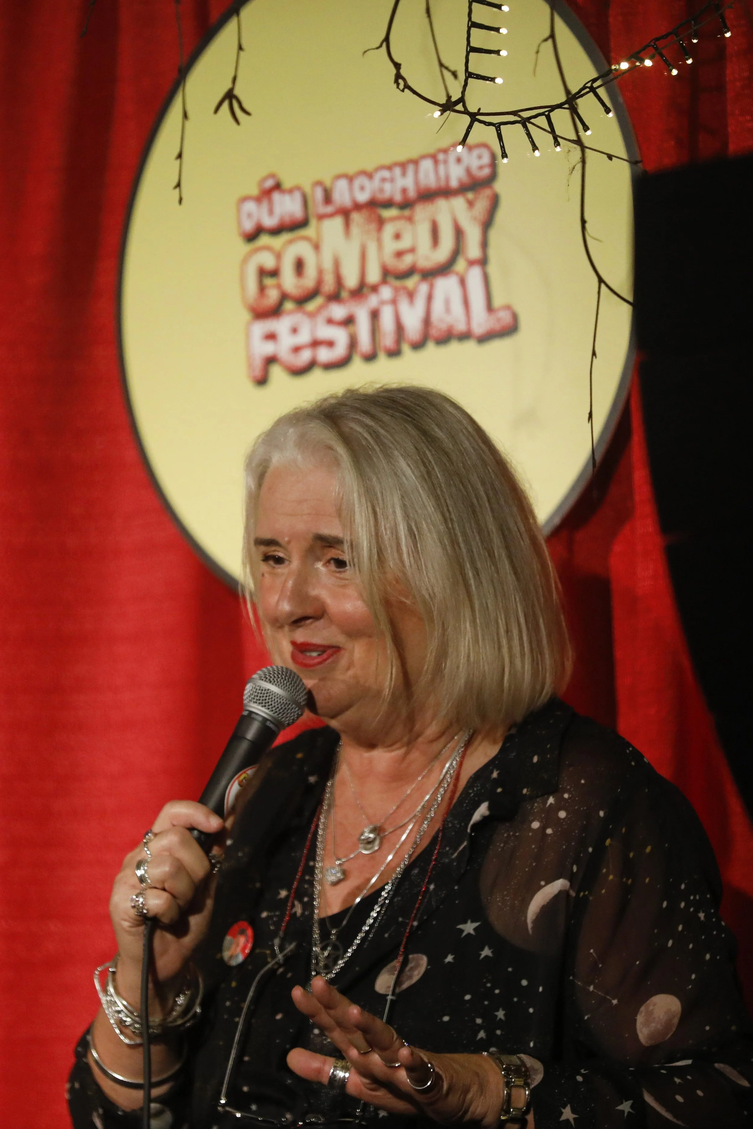 A woman with shoulder-length blond hair speaking into a microphone on stage at the Dún Laoghaire Comedy Festival, with a circular sign in the background reading 'Dún Laoghaire Comedy Festival' and red curtains.