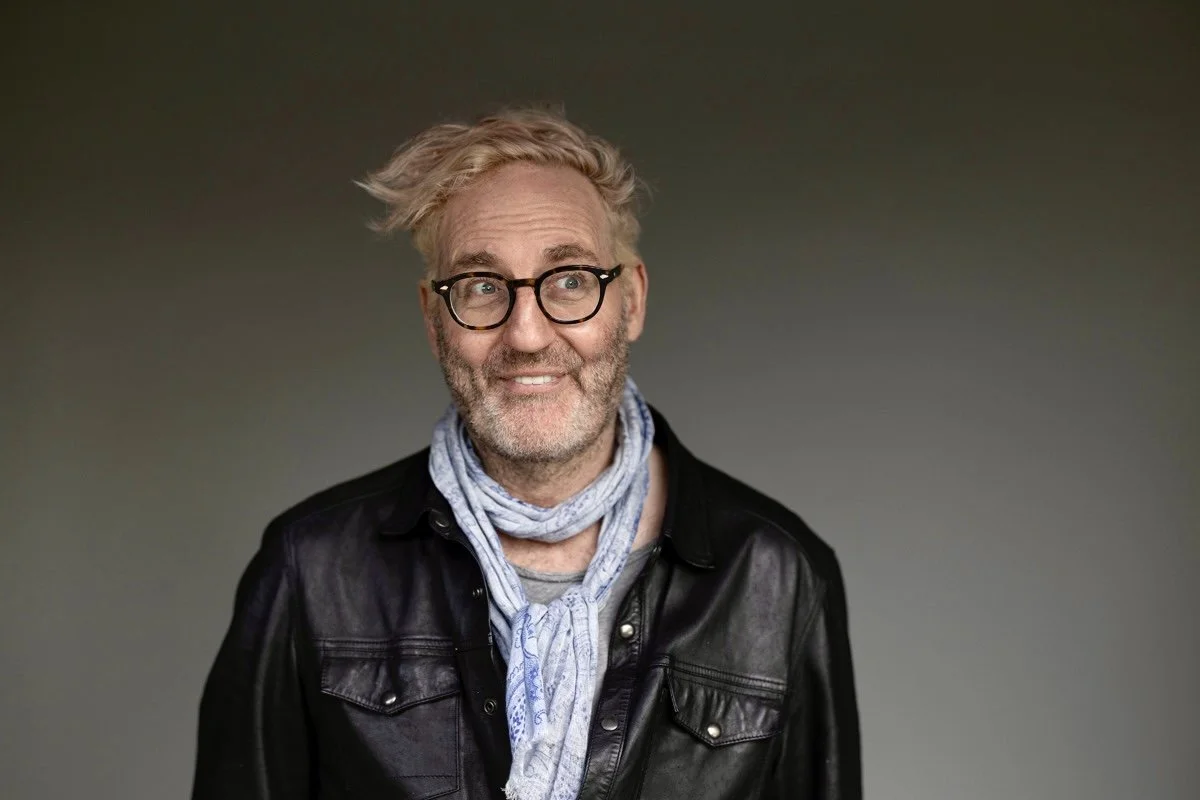 A smiling middle-aged man with wavy, light-colored hair, glasses, a beard, wearing a black leather jacket and a light scarf, standing against a gray background.