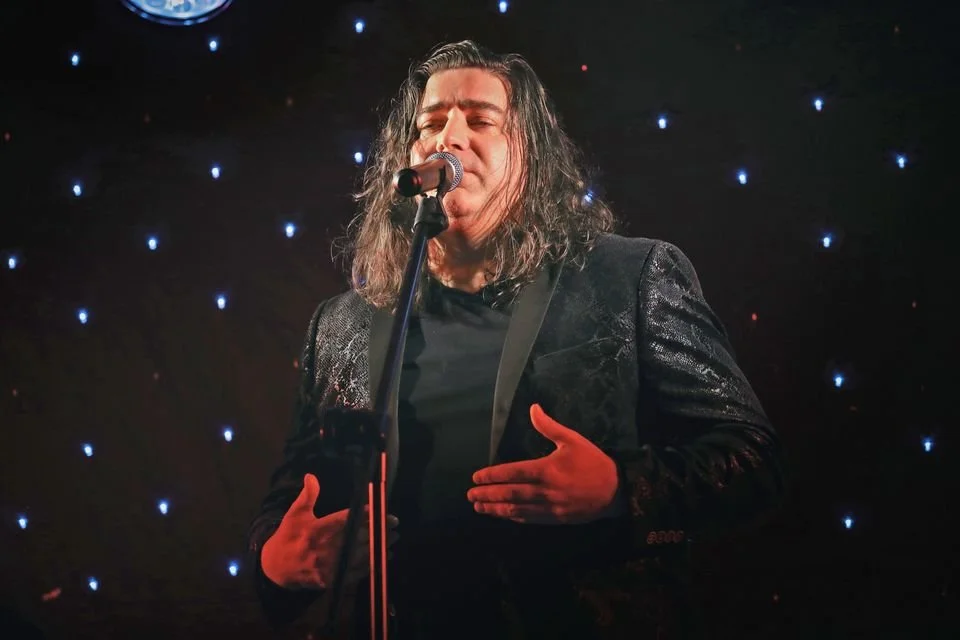 A male singer with long wavy hair performing on stage, singing into a microphone with a hand on his chest, in front of a black starry background.