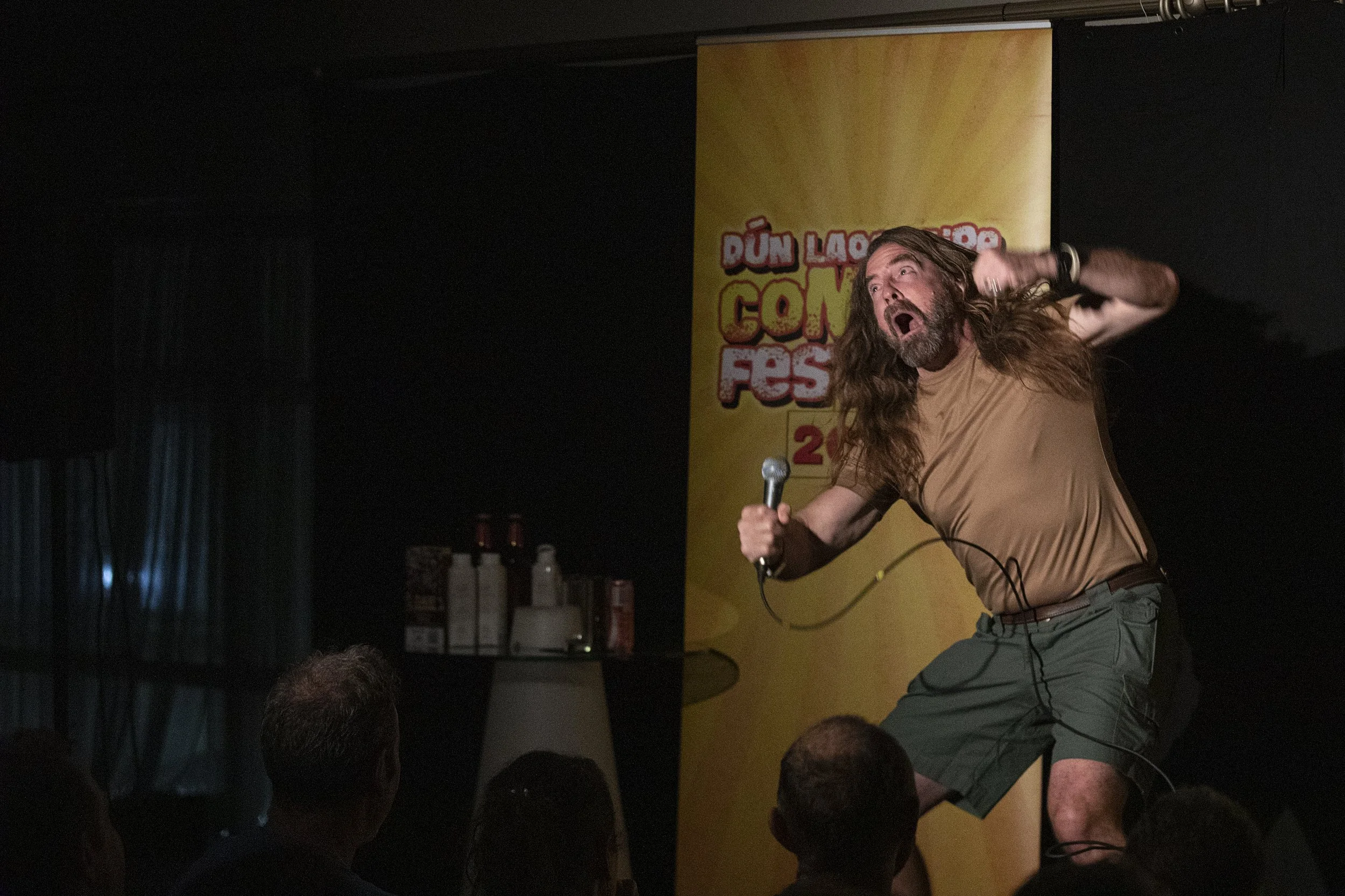 A comedian with long hair and a beard performing on stage at a comedy festival, holding a microphone and making an expressive gesture, with a yellow festival banner in the background.
