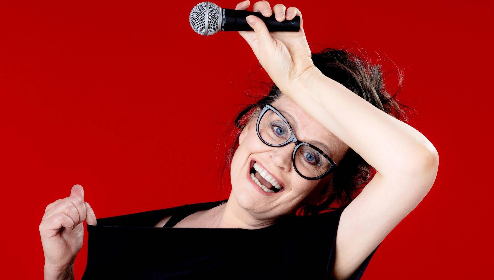 A woman with glasses, dark hair, and a black top, holding a microphone above her head, pulling her shirt to the side, and making a grimace, against a red background.