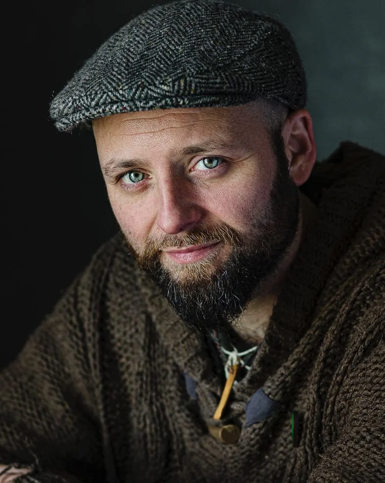 A man with a beard and blue eyes wearing a brown patterned sweater and a gray flat cap.