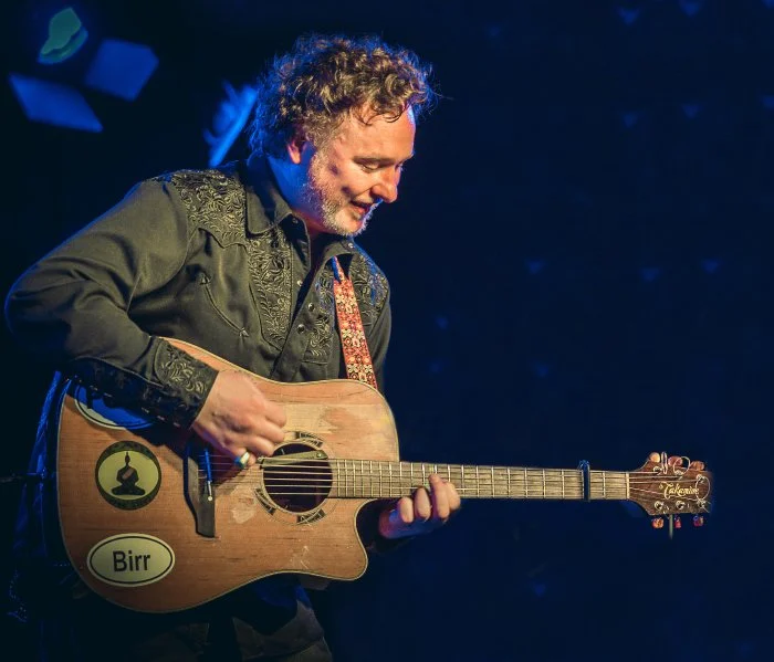 A man with curly hair and a beard playing an acoustic guitar on stage, with a dark blue background and stage lighting.