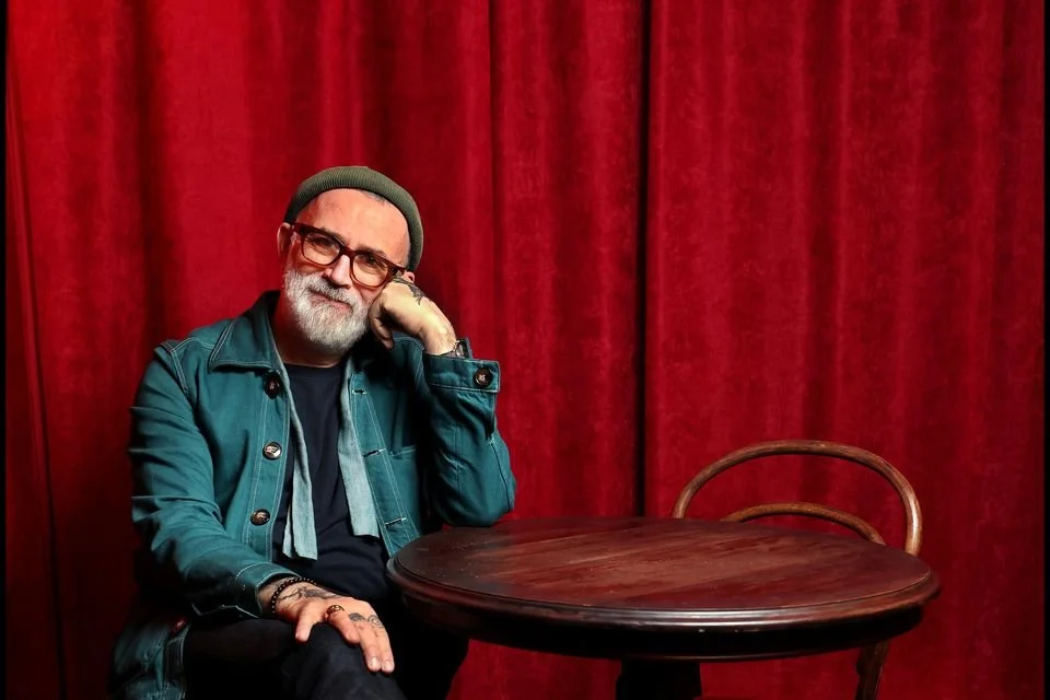 A man with gray hair, glasses, and a beard sitting at a wooden table with a red curtain background.