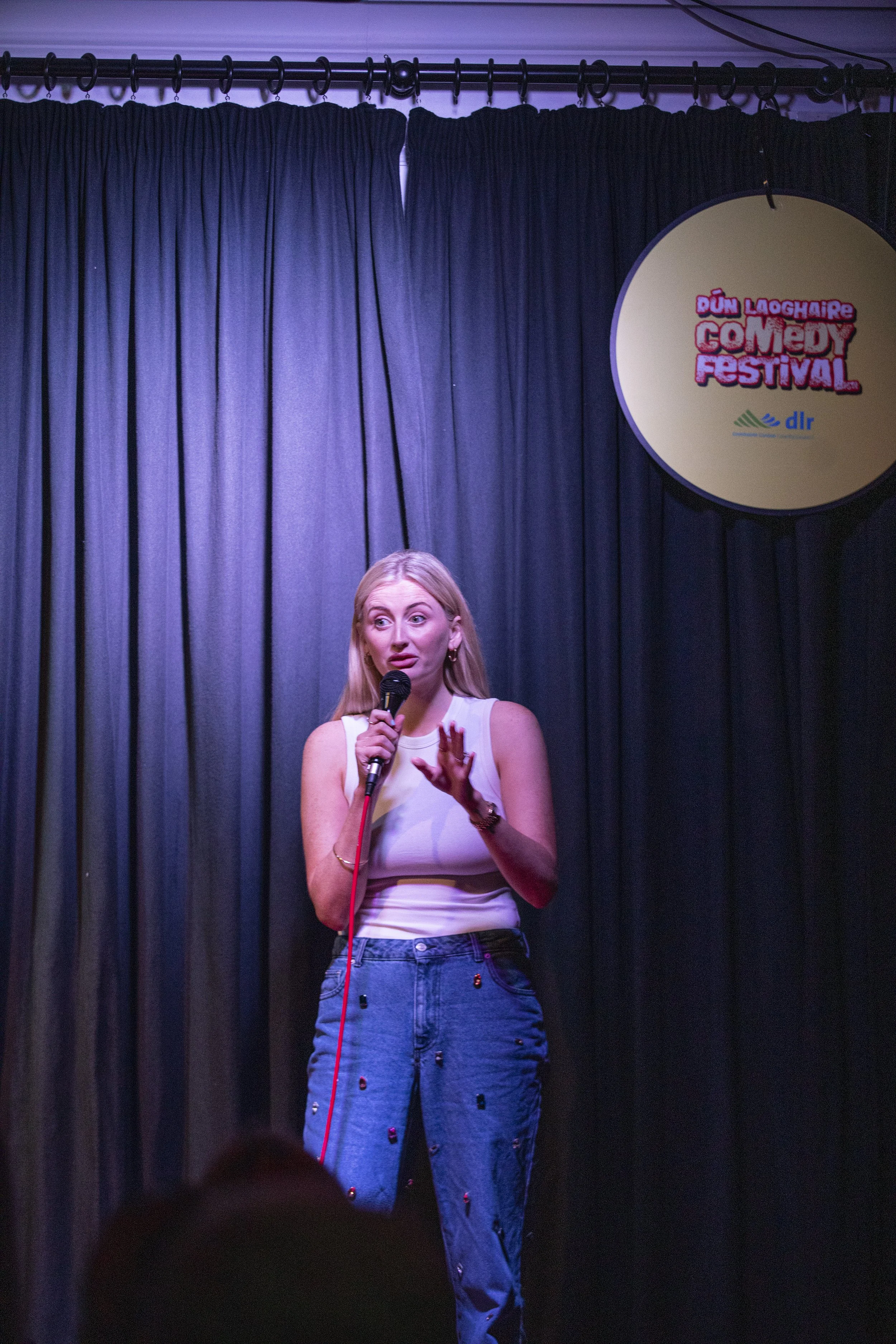 A woman performing stand-up comedy on stage at Dún Laoghaire Comedy Festival, holding a microphone with a dark curtain background.