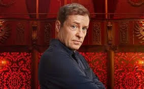 A man with light brown hair, wearing a dark shirt, standing in front of a red ornate carved wooden background.