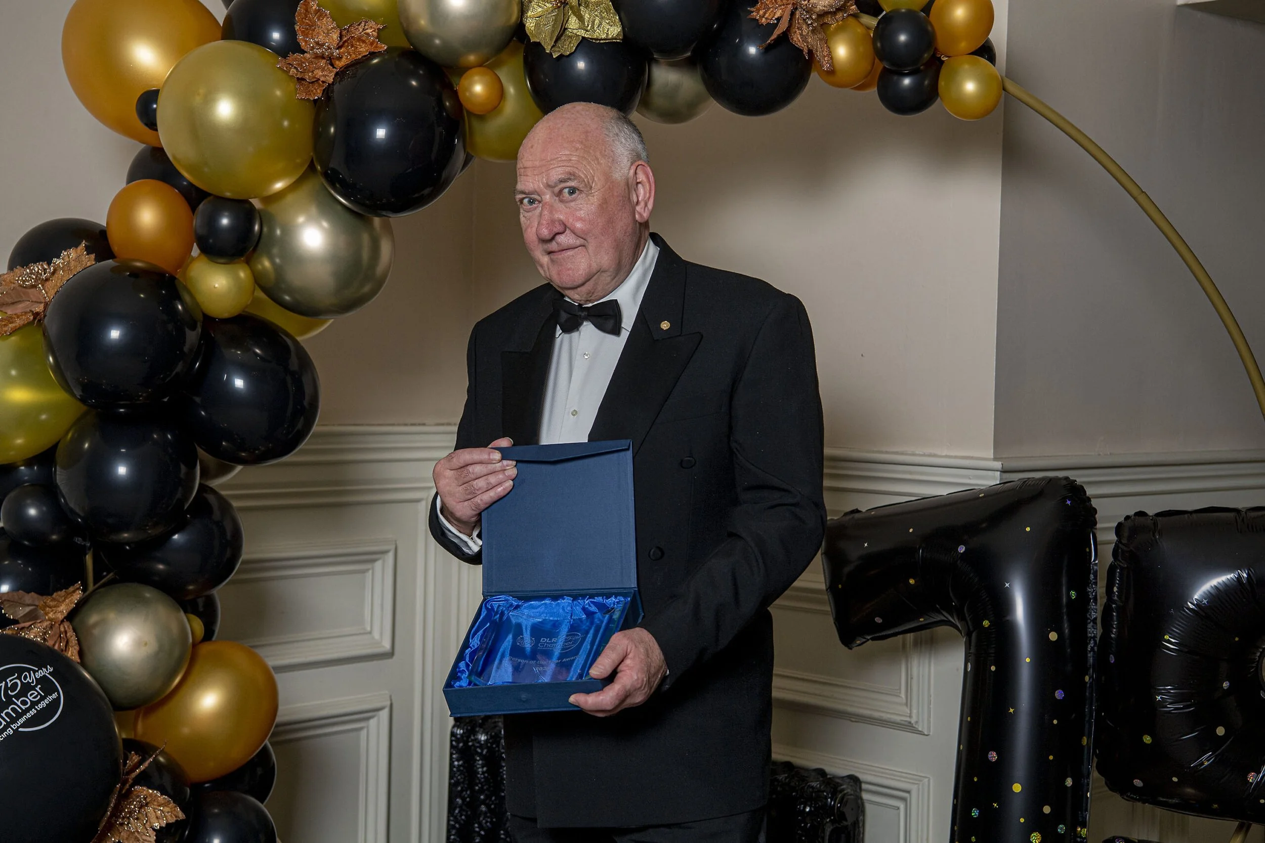 An elderly man in a tuxedo holding an open blue box, standing in front of a balloon arch in black, gold, and green, at a celebration or event.