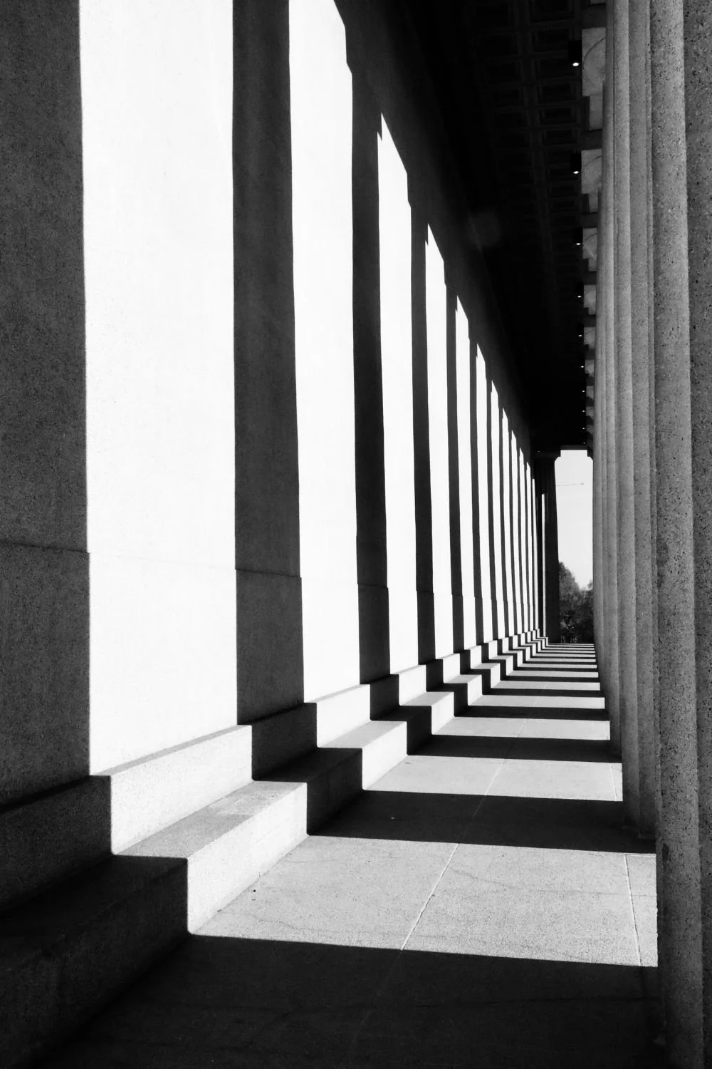 Black and white photo of a colonnade with tall columns casting long shadows on the ground.