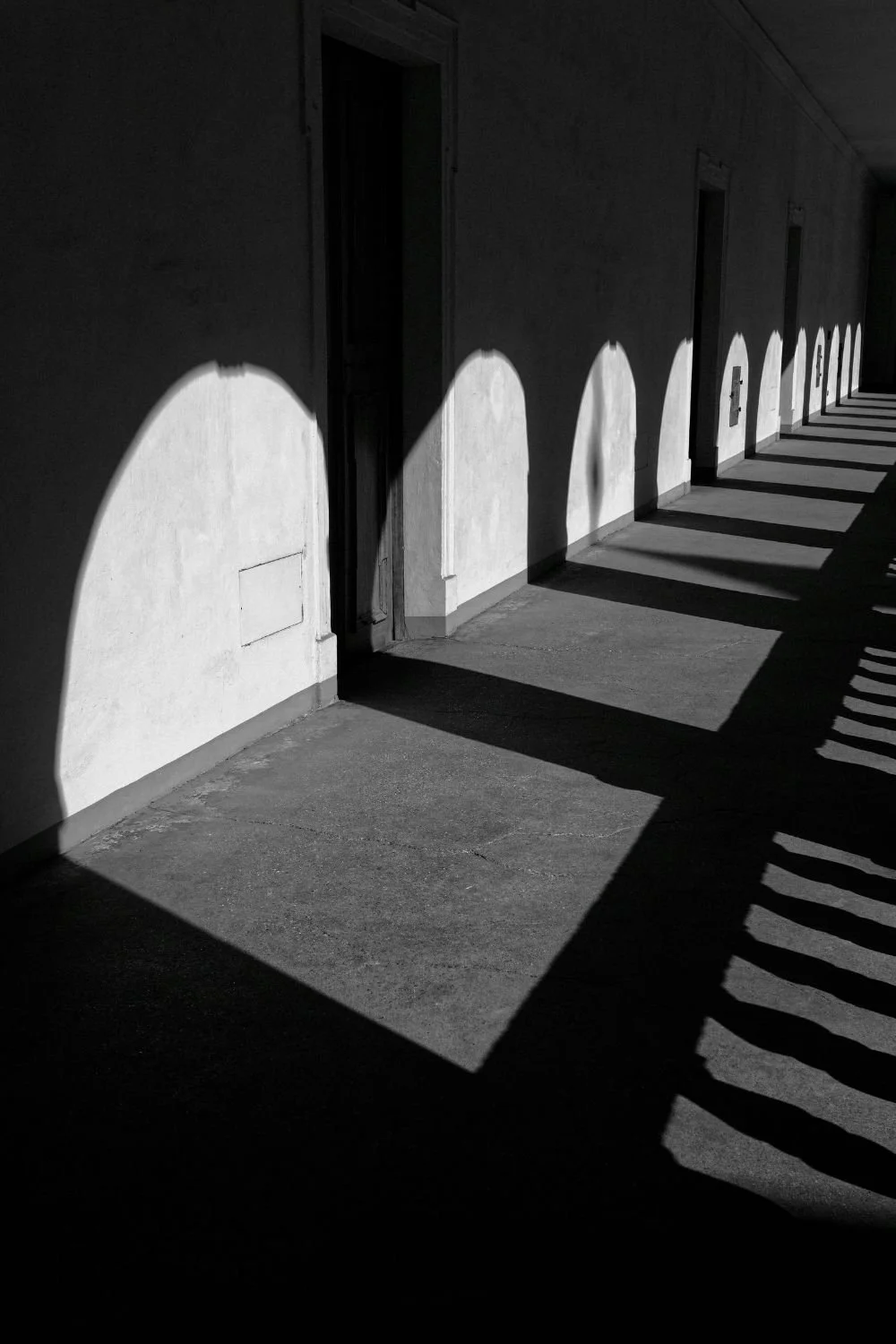 Black and white photo of a building's exterior wall with arched shadows cast by the building's roof overhang, lining along the sidewalk.
