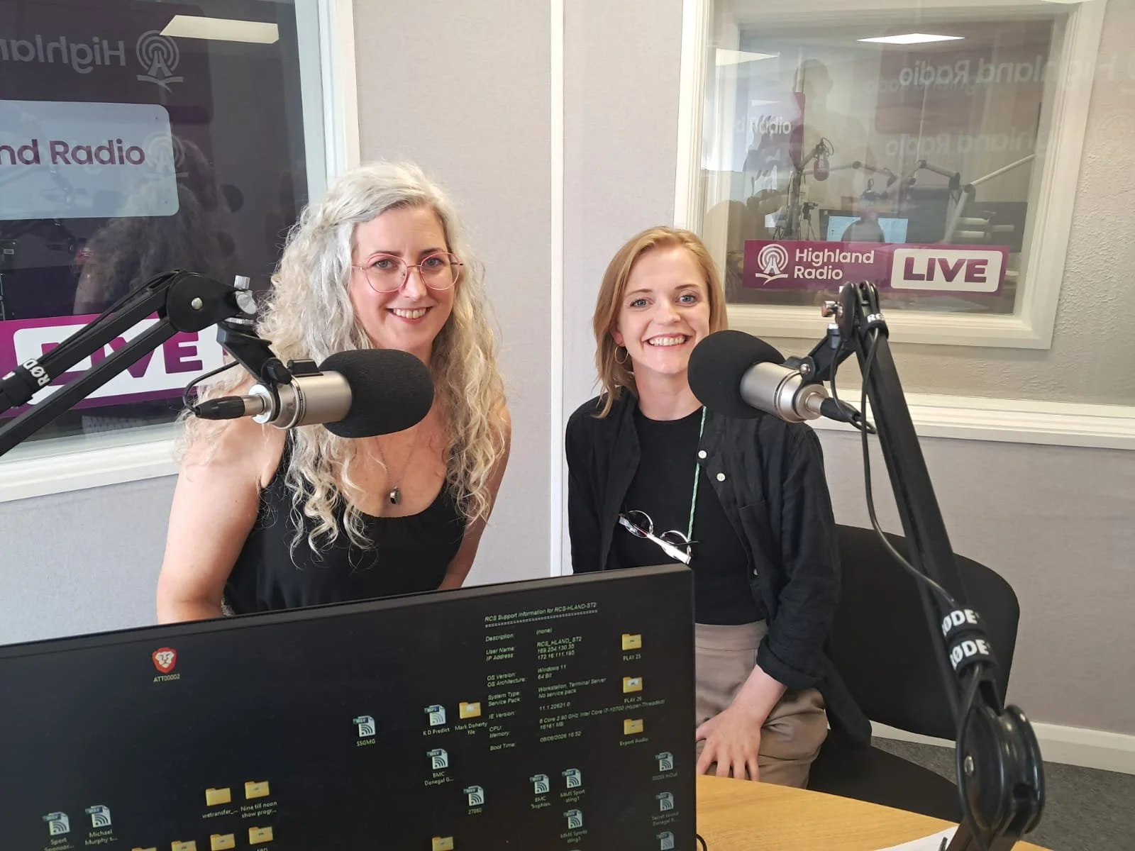 Two women in a radio studio, smiling in front of microphones, with Highland Radio signage visible through a window behind them.