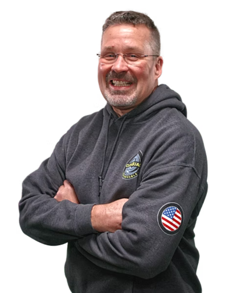 A smiling middle-aged man with glasses and gray hair, wearing a dark gray hoodie with a US flag patch on the sleeve, standing with arms crossed.