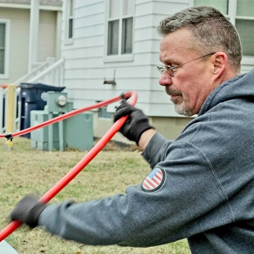 Technician operating drain cleaning cable outside residential home near foundation.
