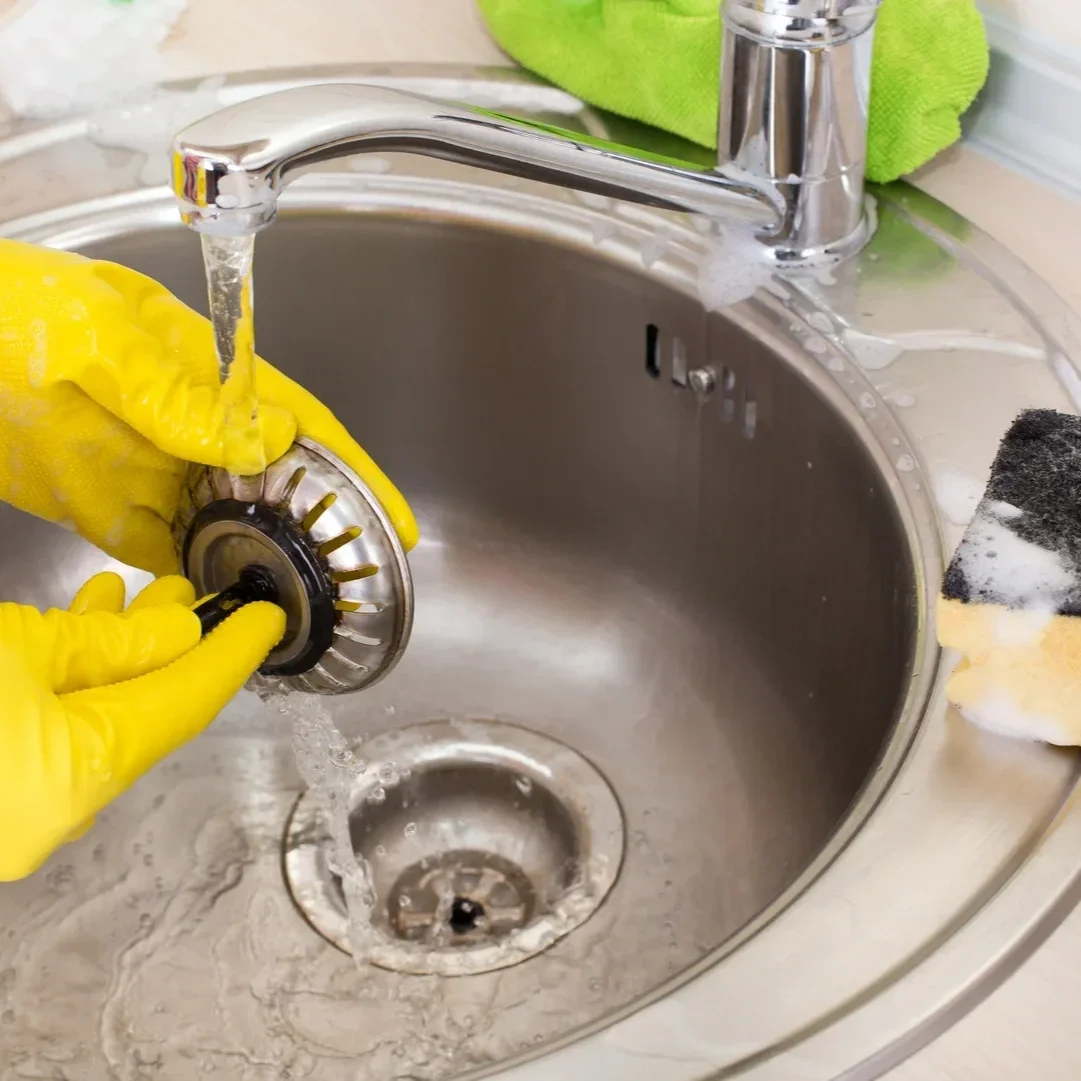 Hands clean a sink drain strainer under running water, illustrating maintenance that removes debris near the drain opening while deeper pipe buildup can still cause slow drainage after snaking.