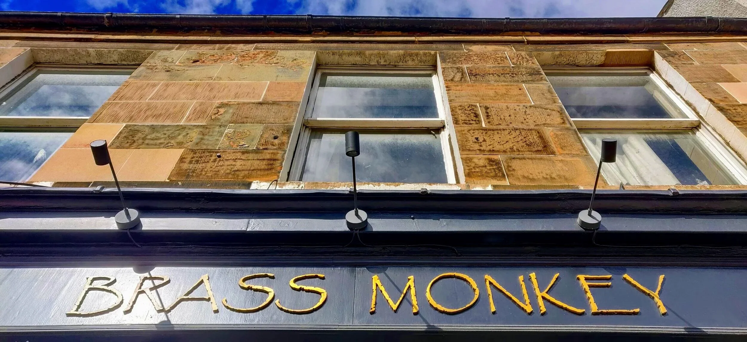 The photograph shows a close-up view of a building's upper facade with brick and stonework. There are three rectangular windows, and hanging from a black architectural ledge below the windows are three small black lamps. Below the lamps, there is a sign with the text 'BASS MONKEY' in yellow and white letters.