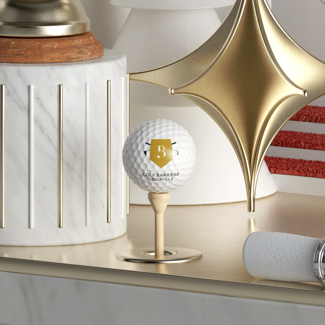 Une boule de golf portant le logo du Golf Barrière Deauville, posée sur un support en bois, entourée d'objets décoratifs sophistiqués en or et en blanc sur une surface beige.