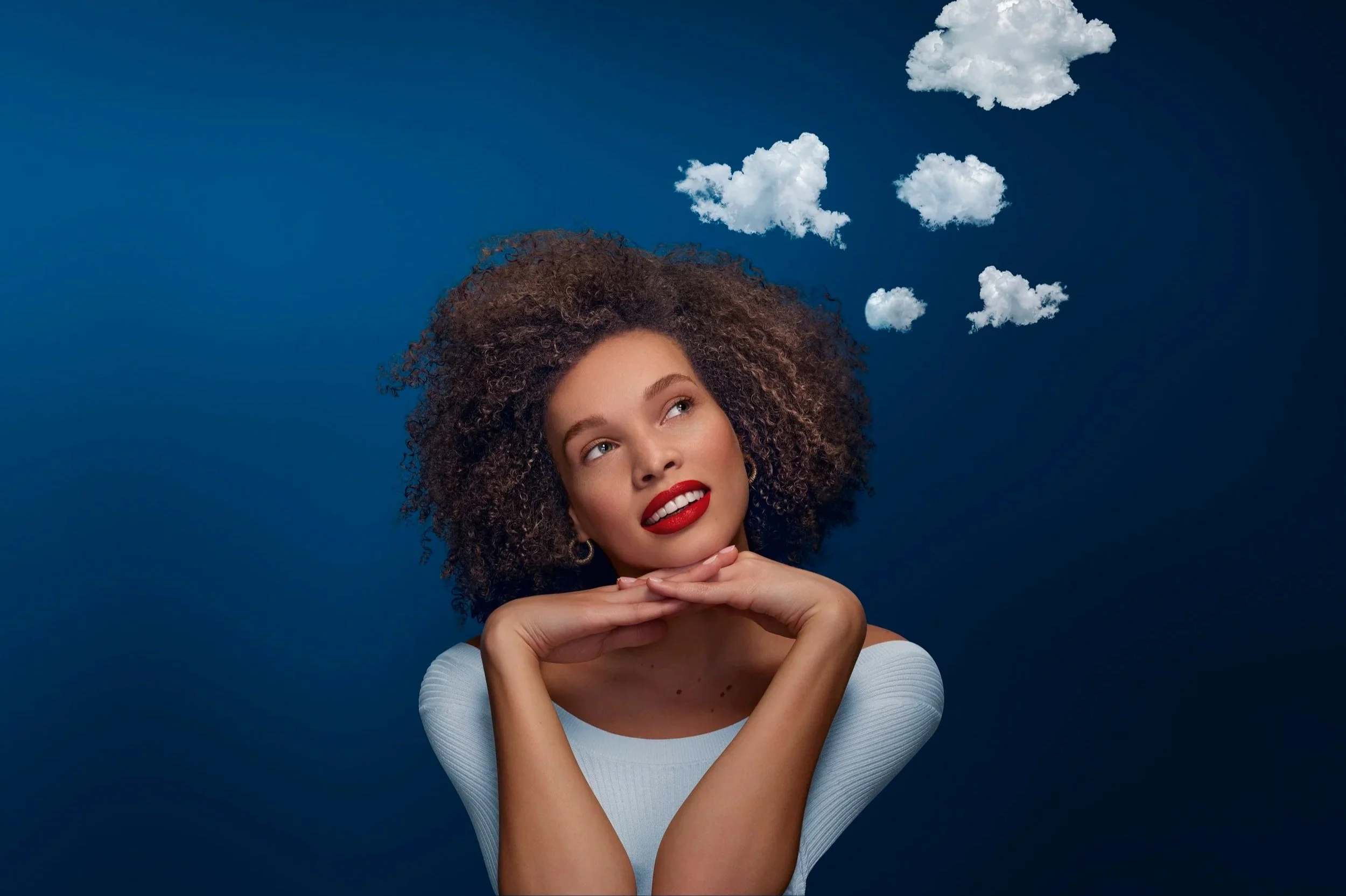 Jeune femme avec cheveux bouclés, souriante en regardant vers le haut, avec des nuages en forme de pensées flottant au-dessus de sa tête, contre un fond bleu.