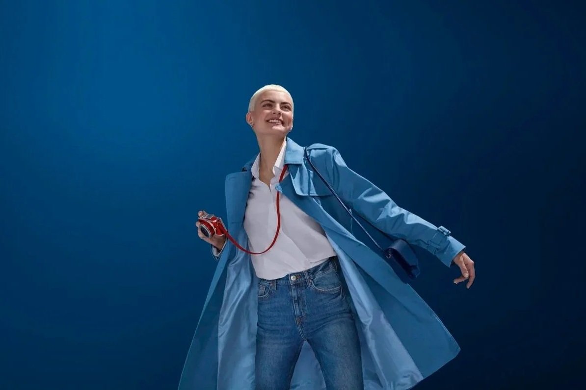 Une femme souriante avec une coiffure courte, portant un long manteau bleu, une chemise blanche, et un jean, tenant une caméra rouge à la main, devant un fond bleu foncé.