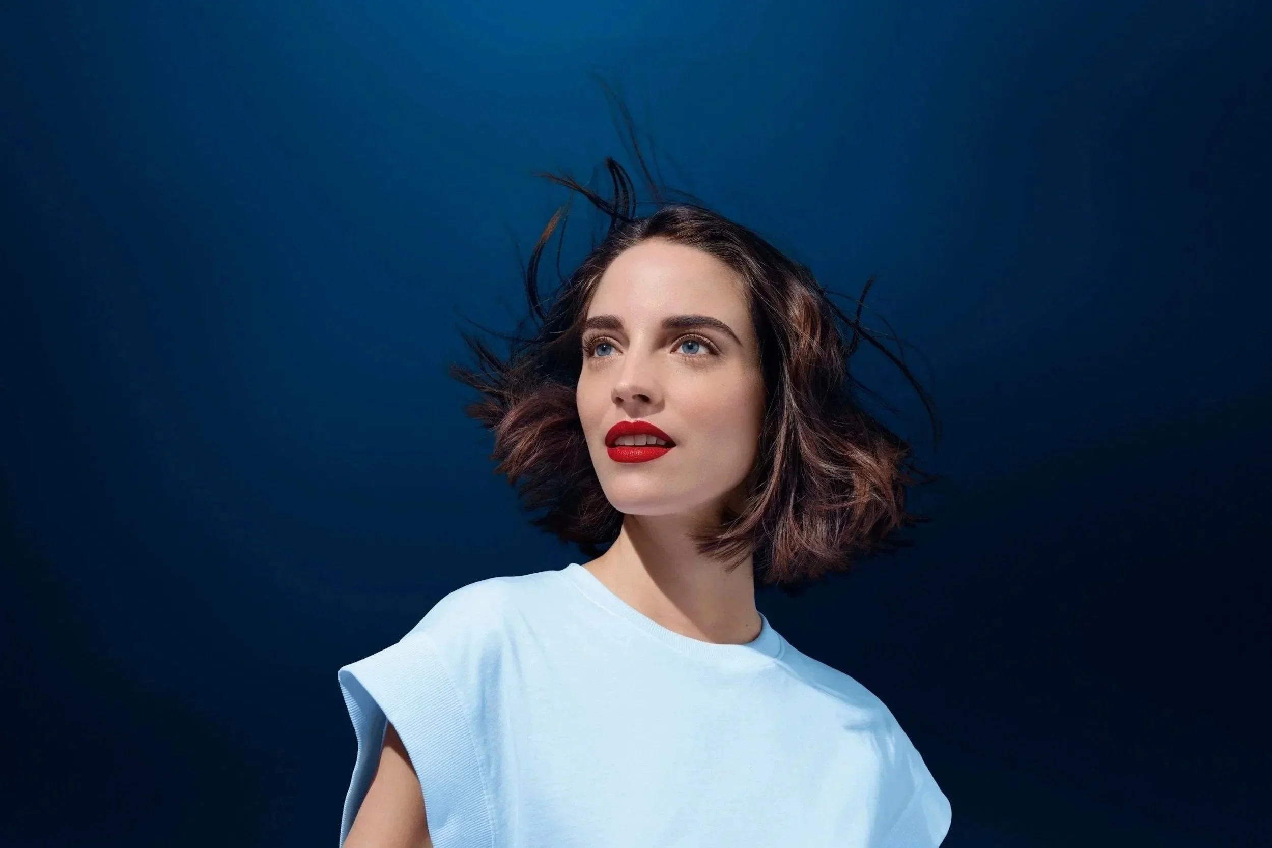 Jeune femme avec cheveux courts en mouvement, portant un t-shirt blanc, regardant de côté, fond bleu sombre.
