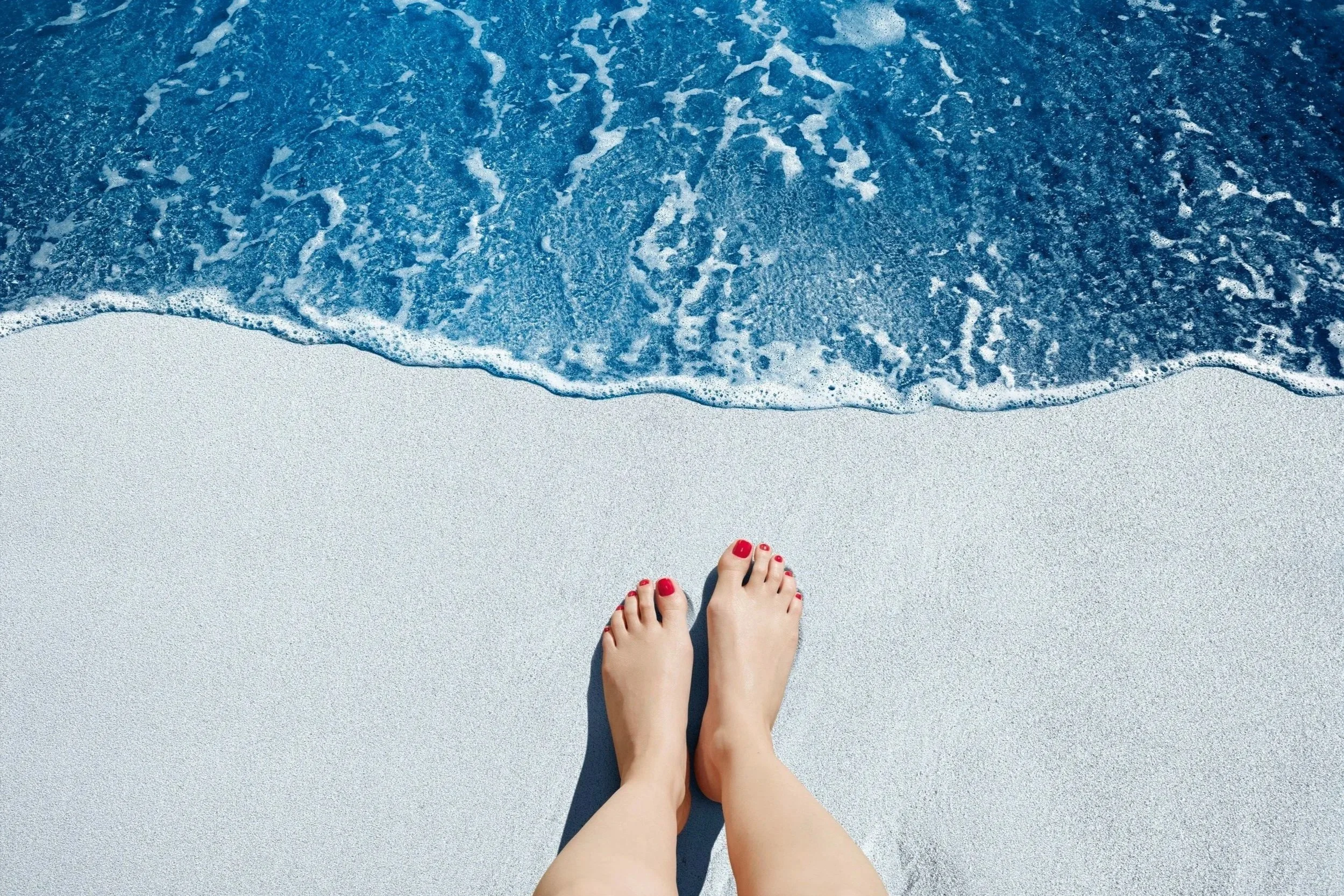 Jambes et pieds d'une personne avec ongles peints en rouge, sur du sable blanc au bord de la mer avec des vagues bleues.