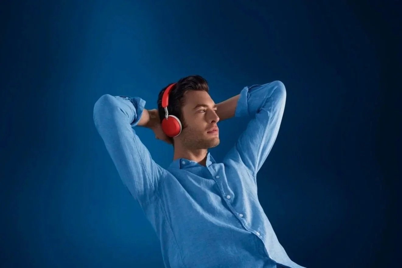 Un jeune homme portant des écouteurs rouges et une chemise bleue, les bras derrière la tête, semblant détendu. Arrière-plan bleu foncé.