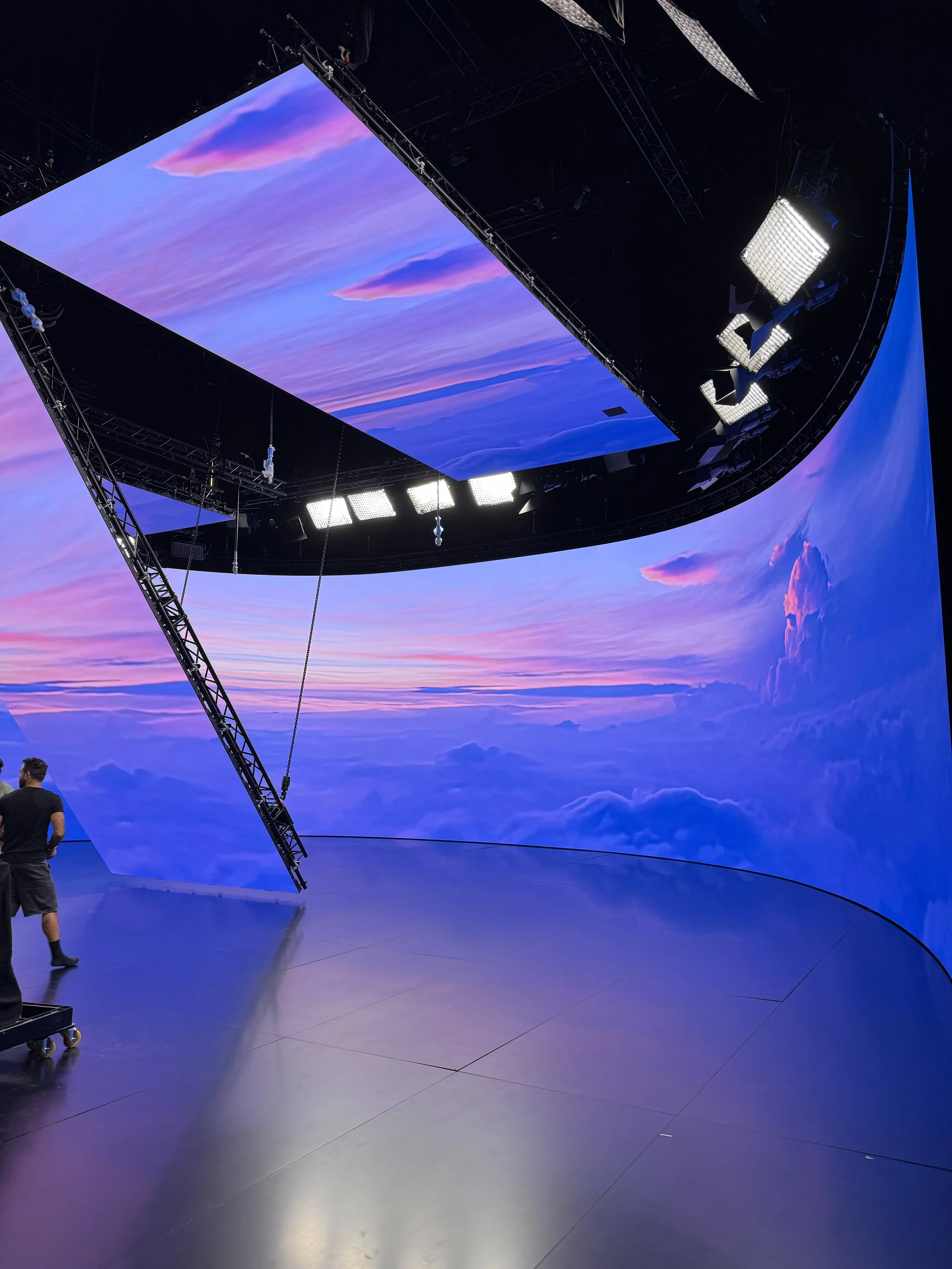 Interior d'une salle avec un grand écran en haut affichant un ciel bleu avec des nuages roses et violets, et un système d'éclairage suspendu au plafond, avec deux personnes dans la pièce.