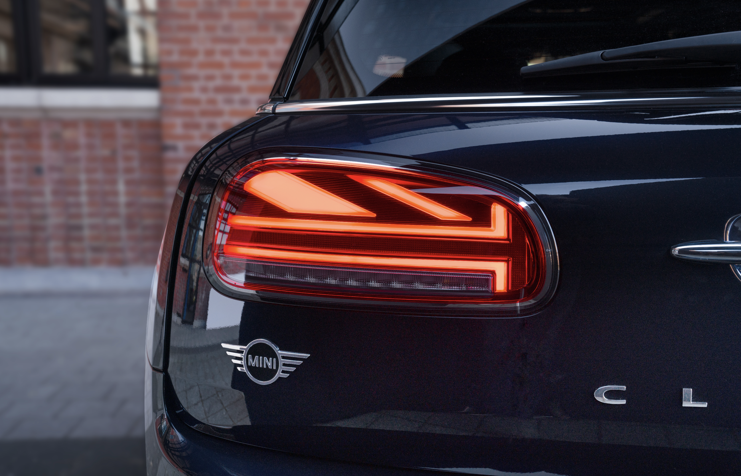 Feux arrière d'une voiture Mini noire, avec logo Mini et lettres 'CL', stationnée devant un mur en brique.