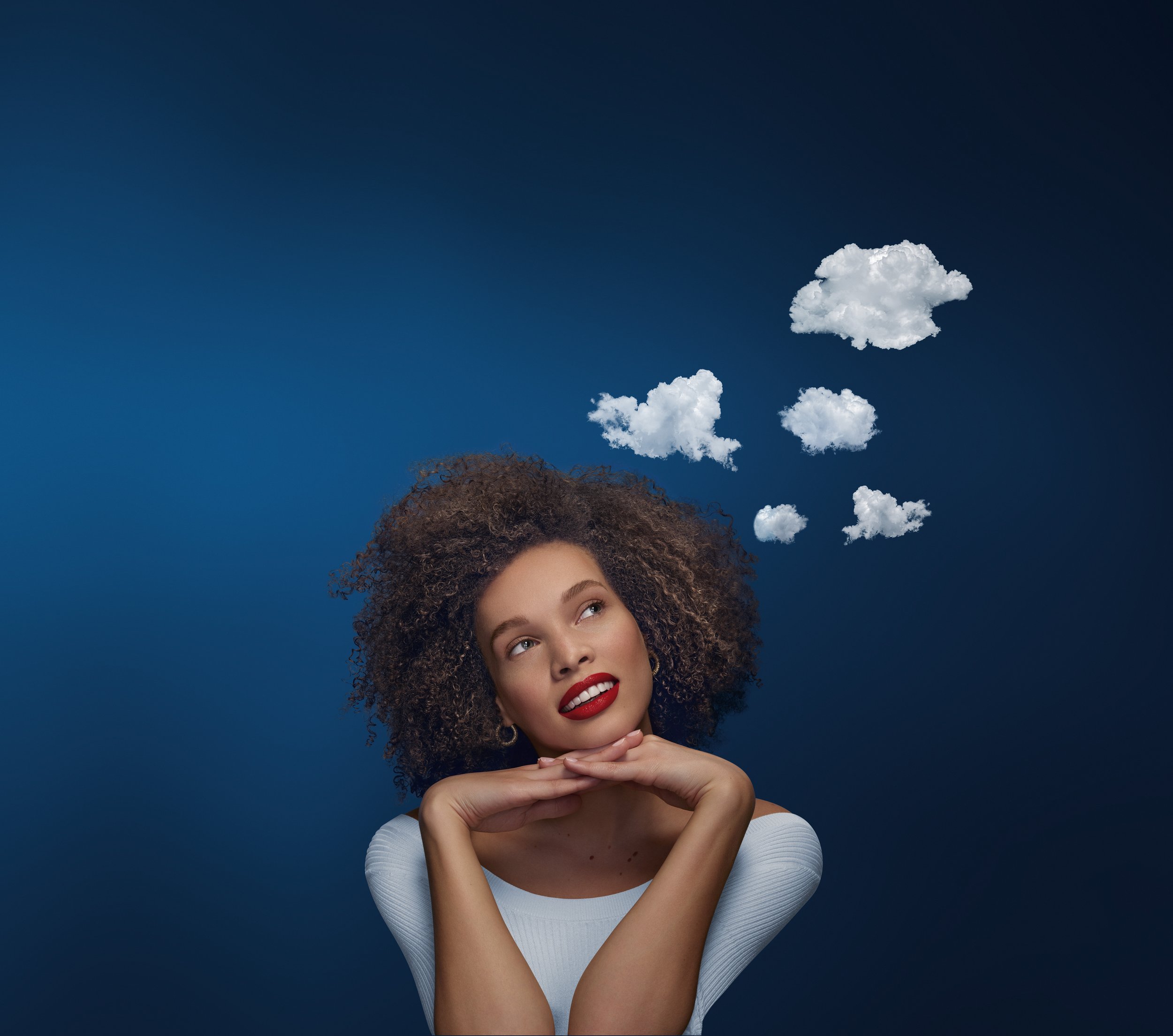 Jeune femme avec cheveux bouclés, souriante en regardant vers le haut, avec des nuages en forme de pensées flottant au-dessus de sa tête, contre un fond bleu.