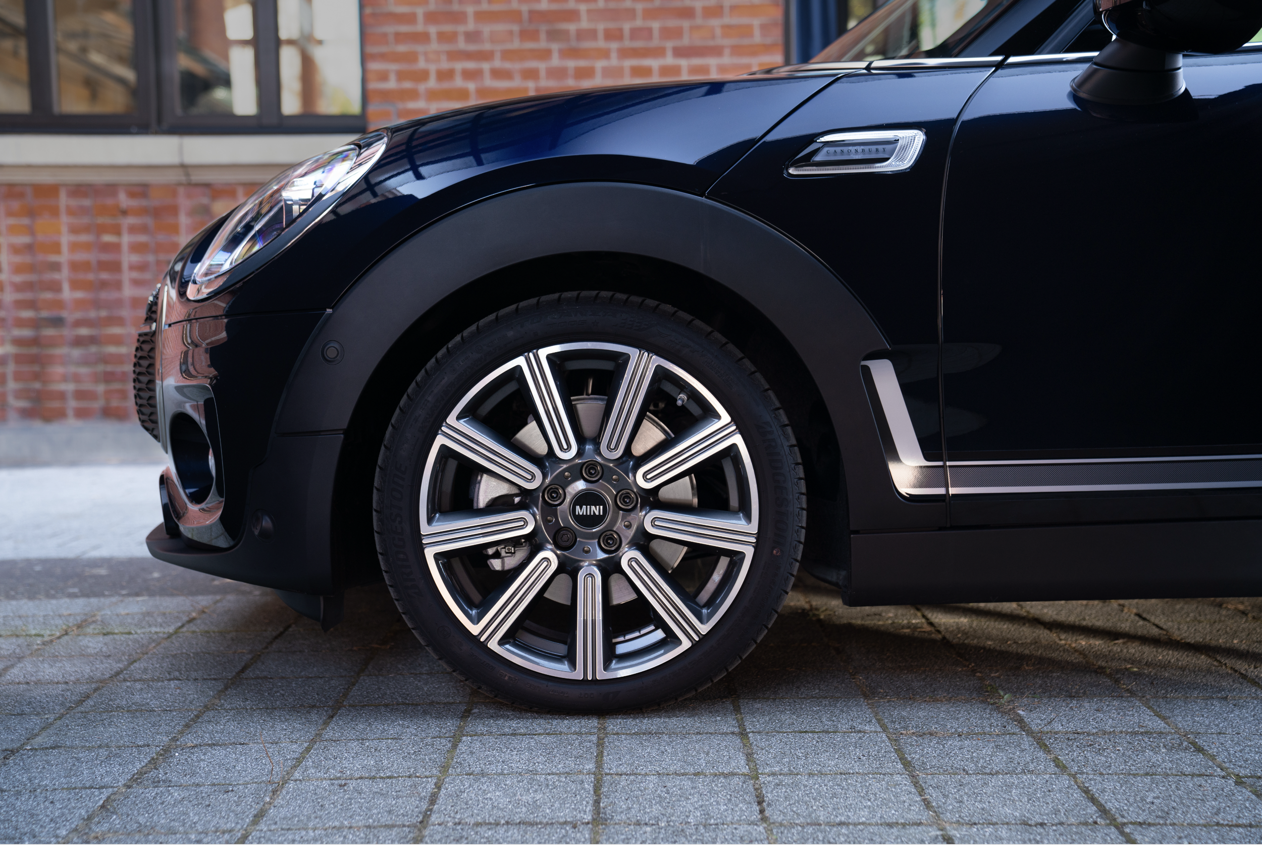 Une voiture noire MINI avec des jantes en alliage circulaires, stationnée sur un pavé devant un bâtiment en briques.