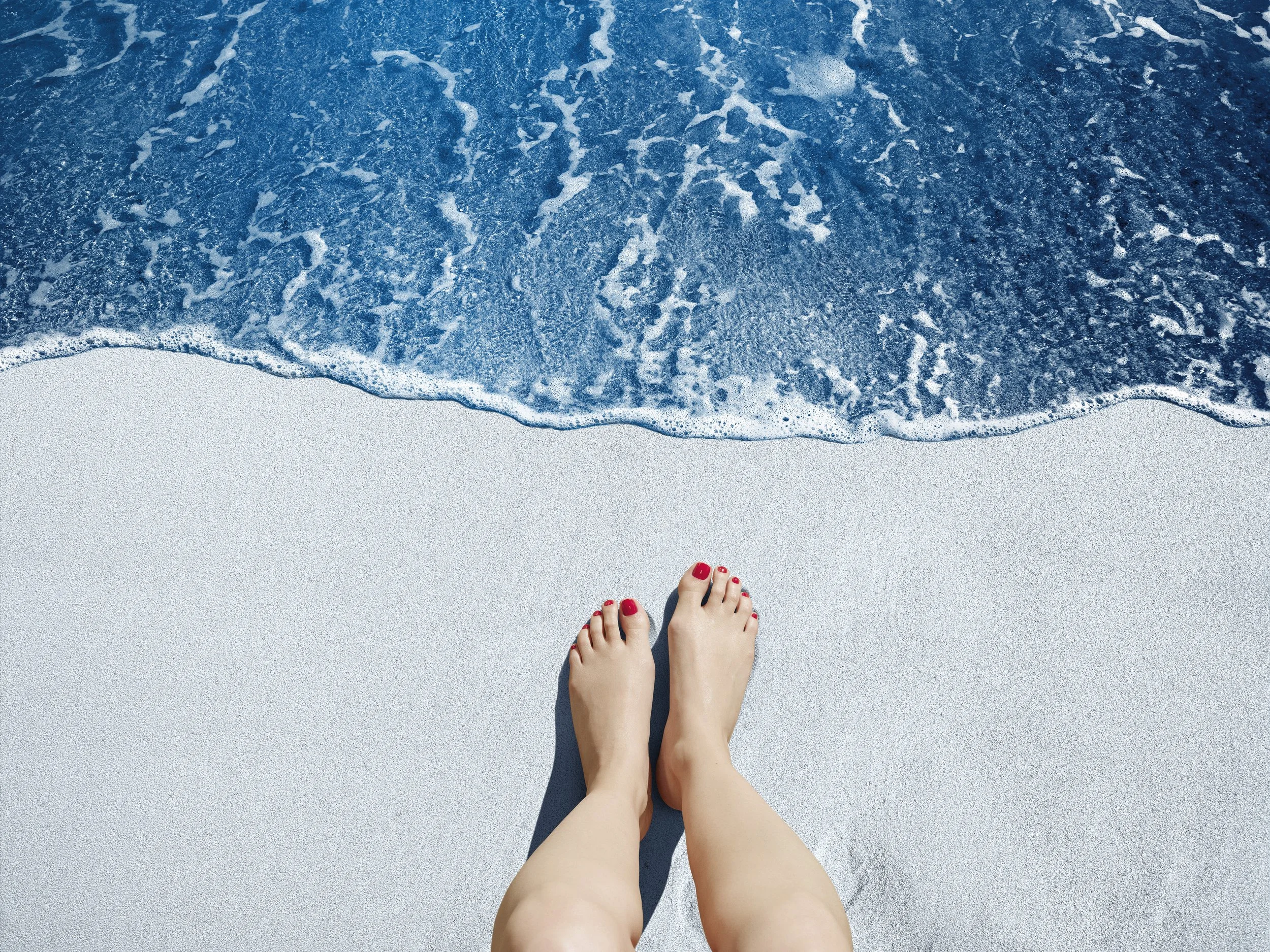 Jambes et pieds d'une personne avec ongles peints en rouge, sur du sable blanc au bord de la mer avec des vagues bleues.