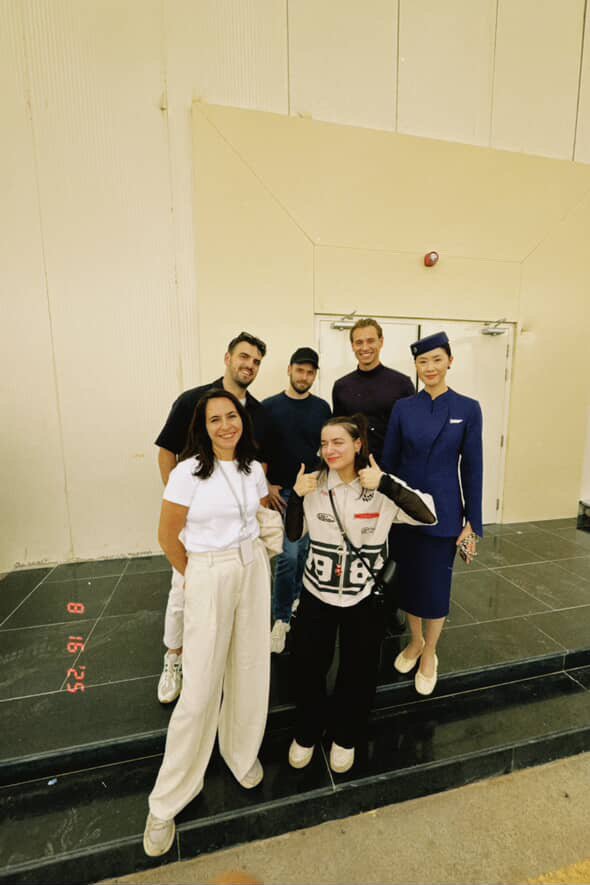 Groupe de six personnes posant pour une photo en intérieur, certains souriants, une femme habillée en uniforme de steward dans le fond.