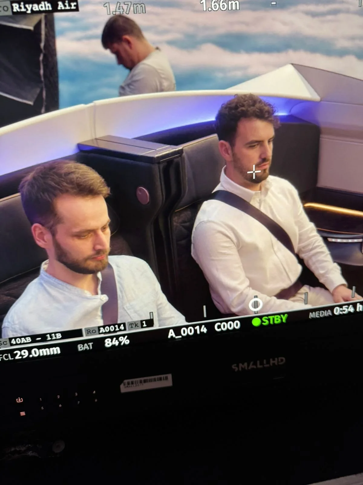 Deux hommes sont assis dans un avion ou une simulation de cockpit, portant des ceintures de sécurité, avec un décor d'arrière-plan de ciel et de nuages. Un troisième homme se tient derrière eux, regardant vers le bas.