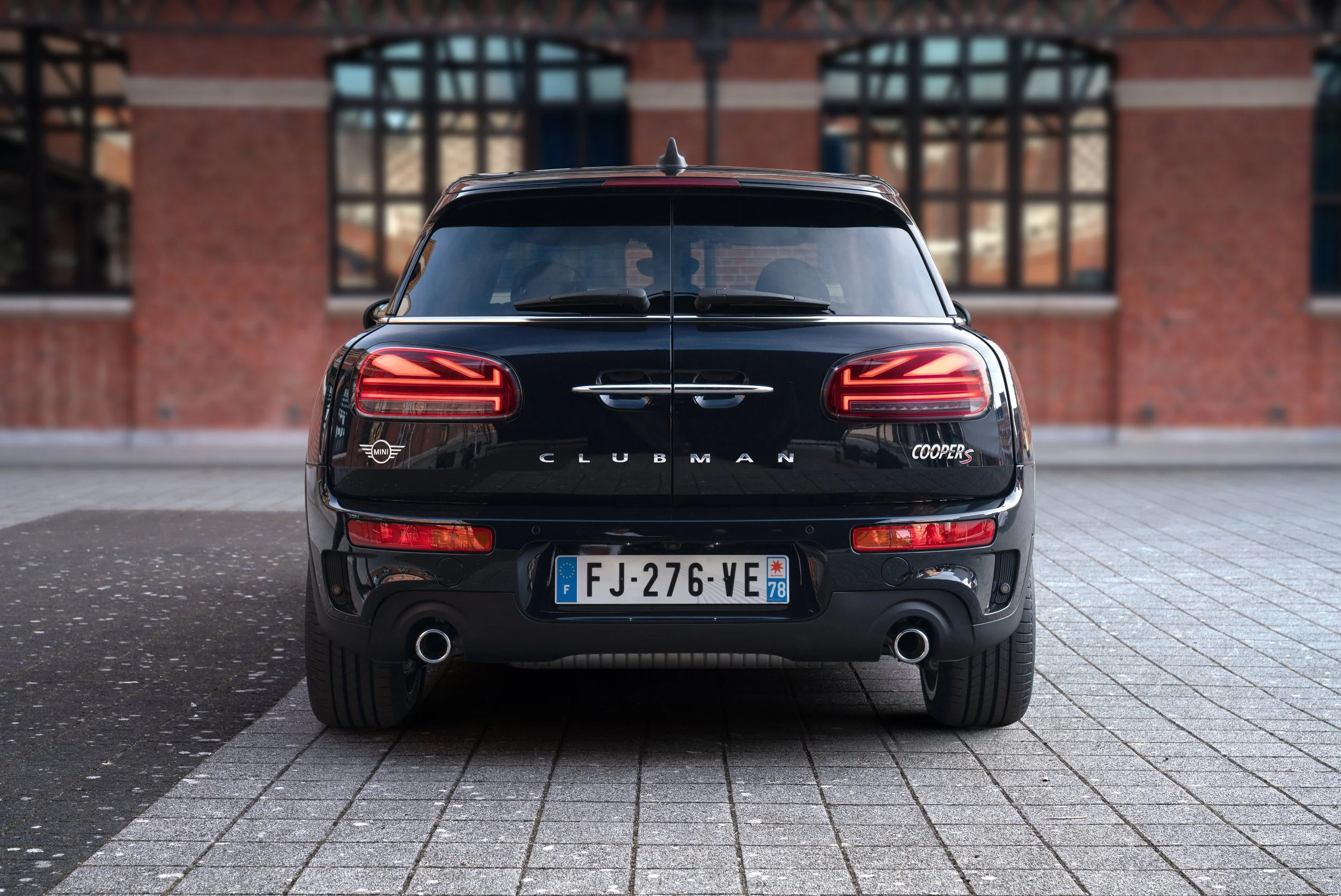 Vue arrière d'une voiture noire Mini Cooper S Clubman, stationnée sur un trottoir, avec un bâtiment en brique en arrière-plan.