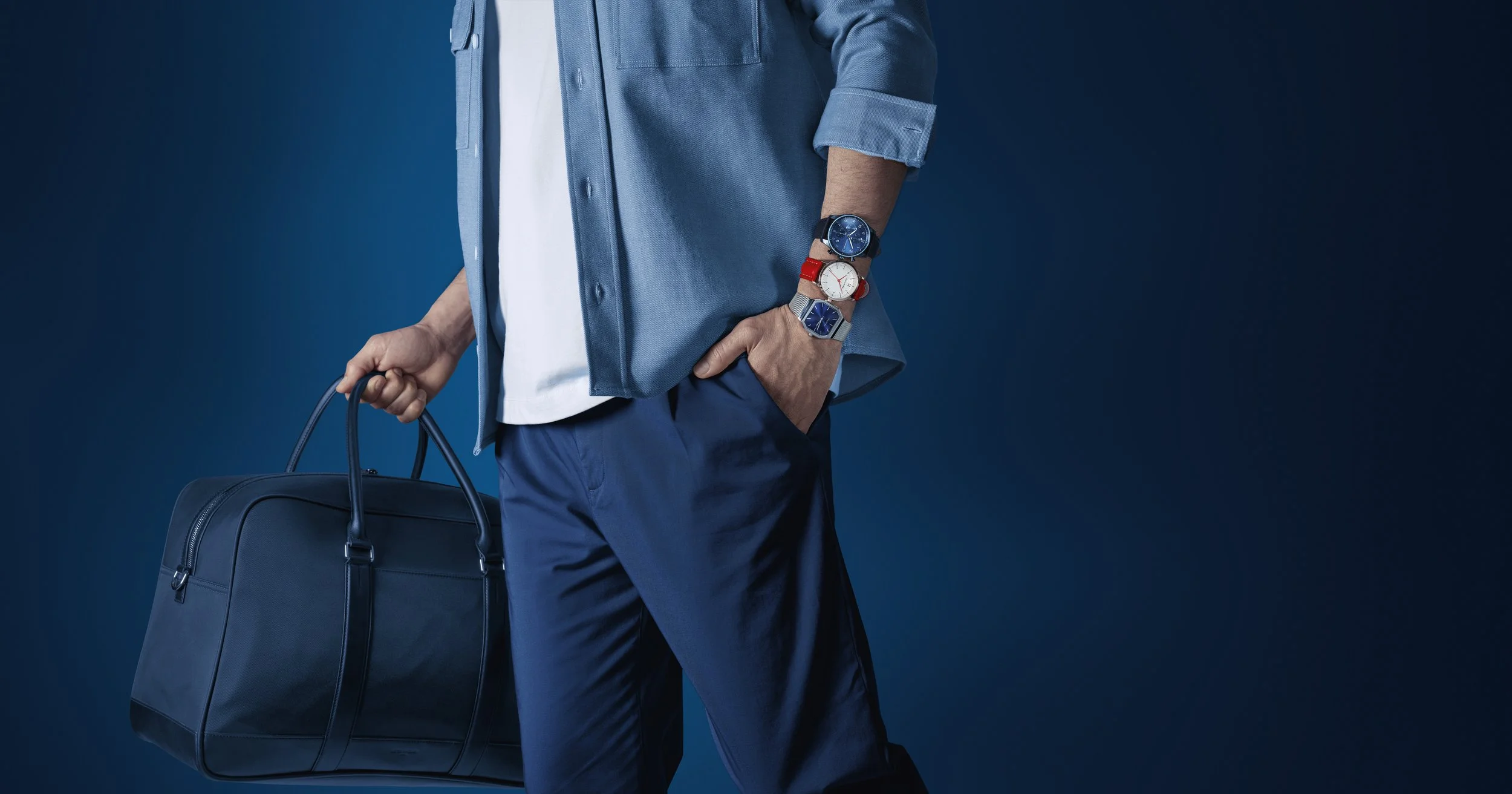 Homme habillé en bleu avec un sac de voyage et plusieurs montres portées au poignet, contre un fond bleu foncé.