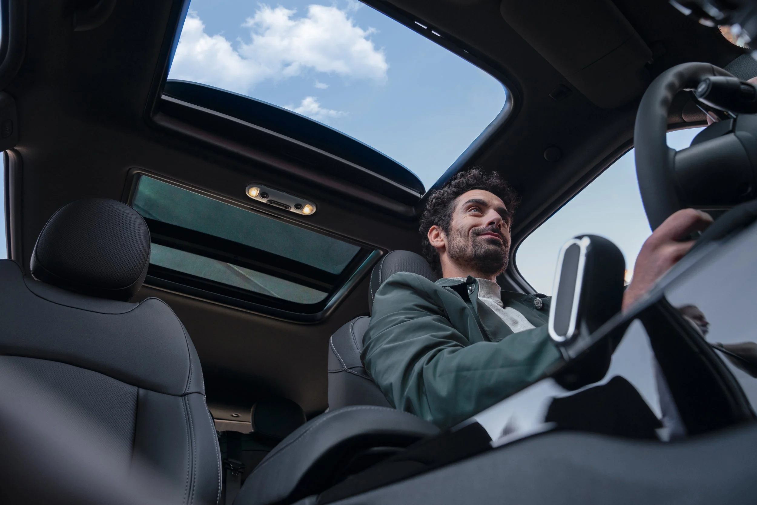 Homme souriant au volant d'une voiture avec toit ouvrant et ciel bleu visible.