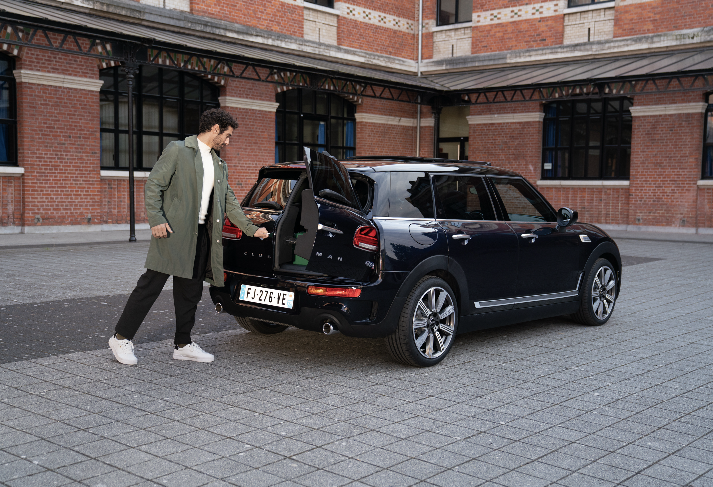 Un homme ouvre le coffre d'une voiture noire en ville, à côté d'un bâtiment en briques rouges, portant une veste et un pantalon noirs, avec des étoles blanches et des baskets blanches.