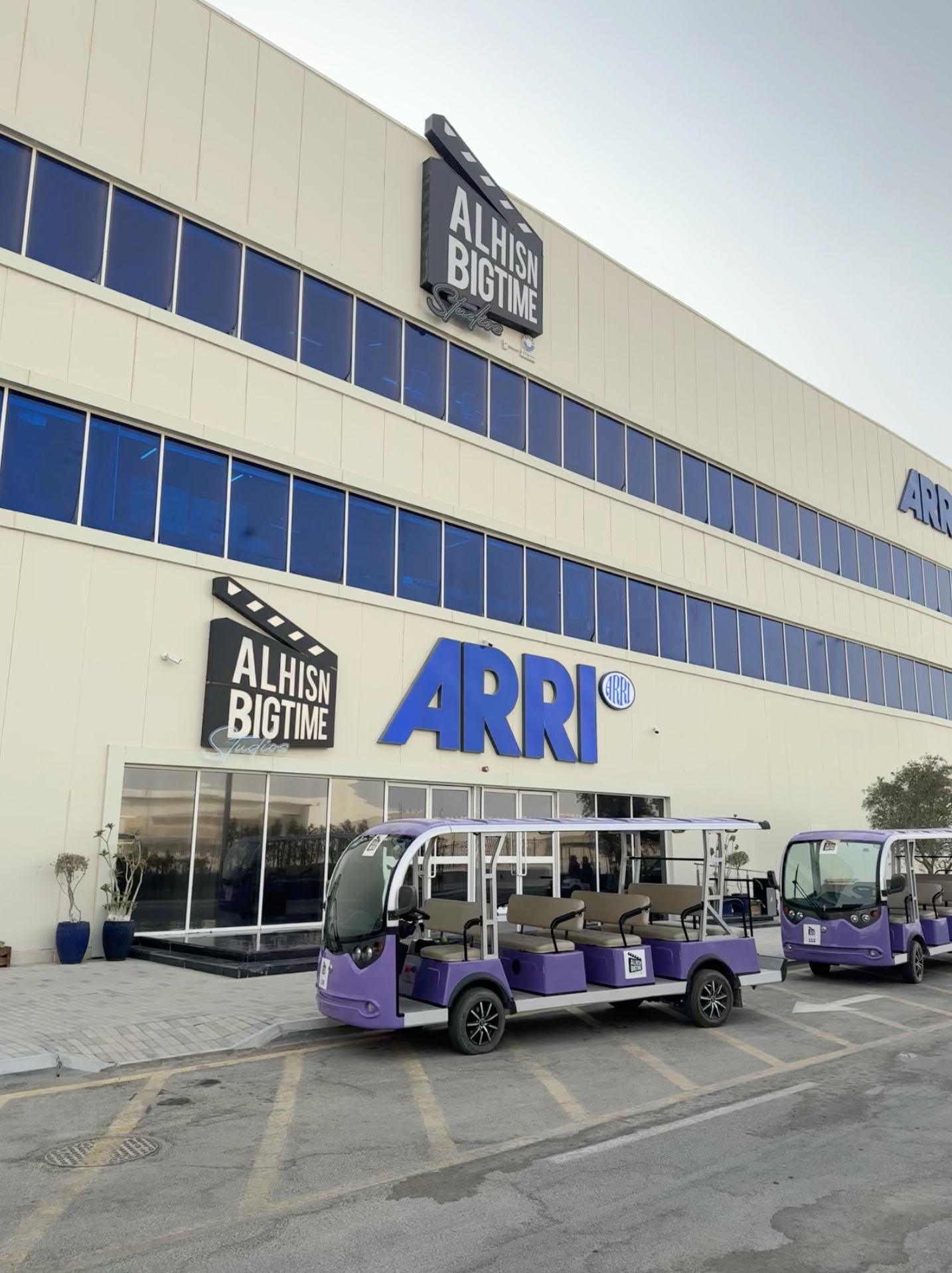 Vue de l'extérieur d'un bâtiment commercial avec le nom 'ARRI' en lettres bleues et un panneau 'Alhisin Bigtime Studios'. Des voitures électriques violettes avec sièges sont garées devant, ainsi qu'une petite structure pour les passagers.