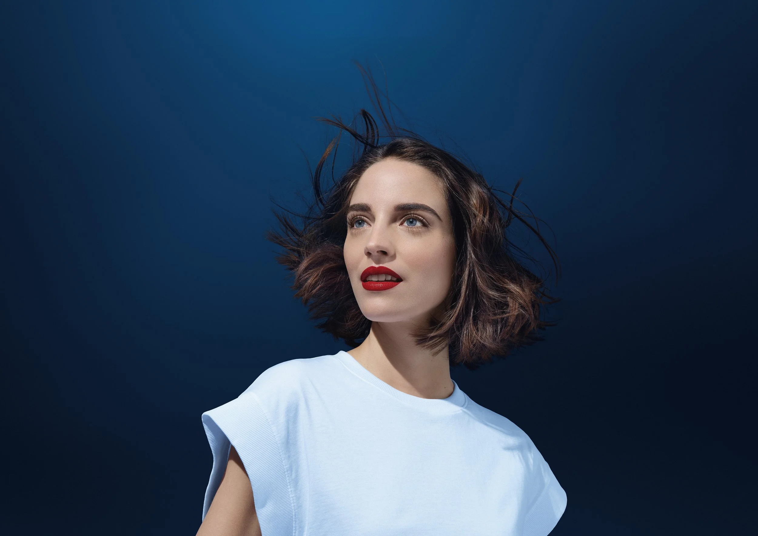 Jeune femme avec cheveux courts en mouvement, portant un t-shirt blanc, regardant de côté, fond bleu sombre.